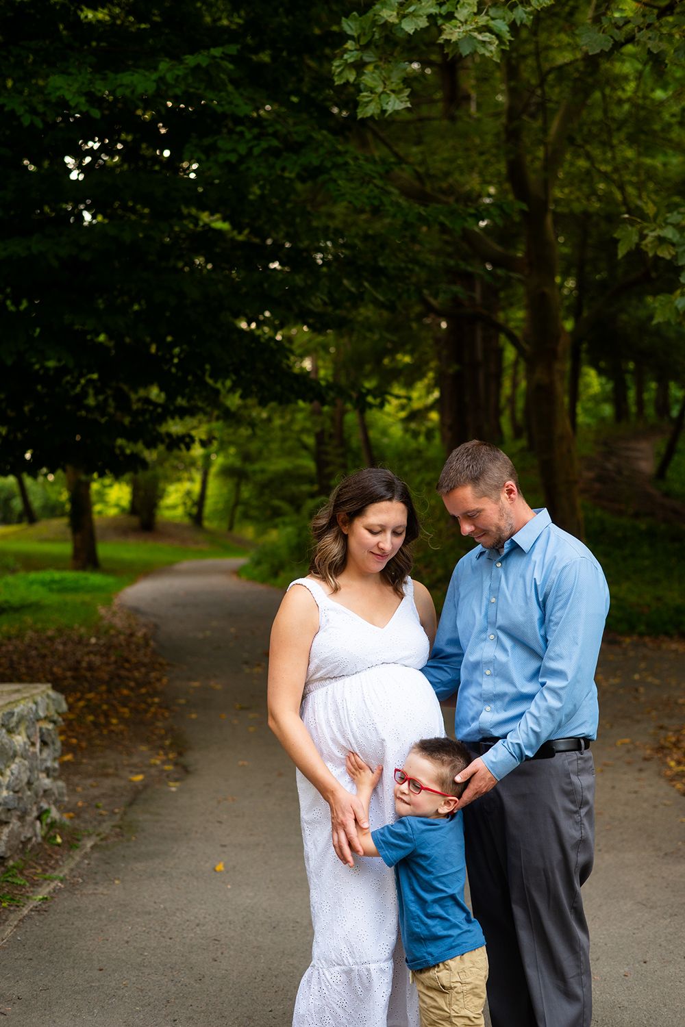 A couple in casual attire stands with their child on a tree-lined path.