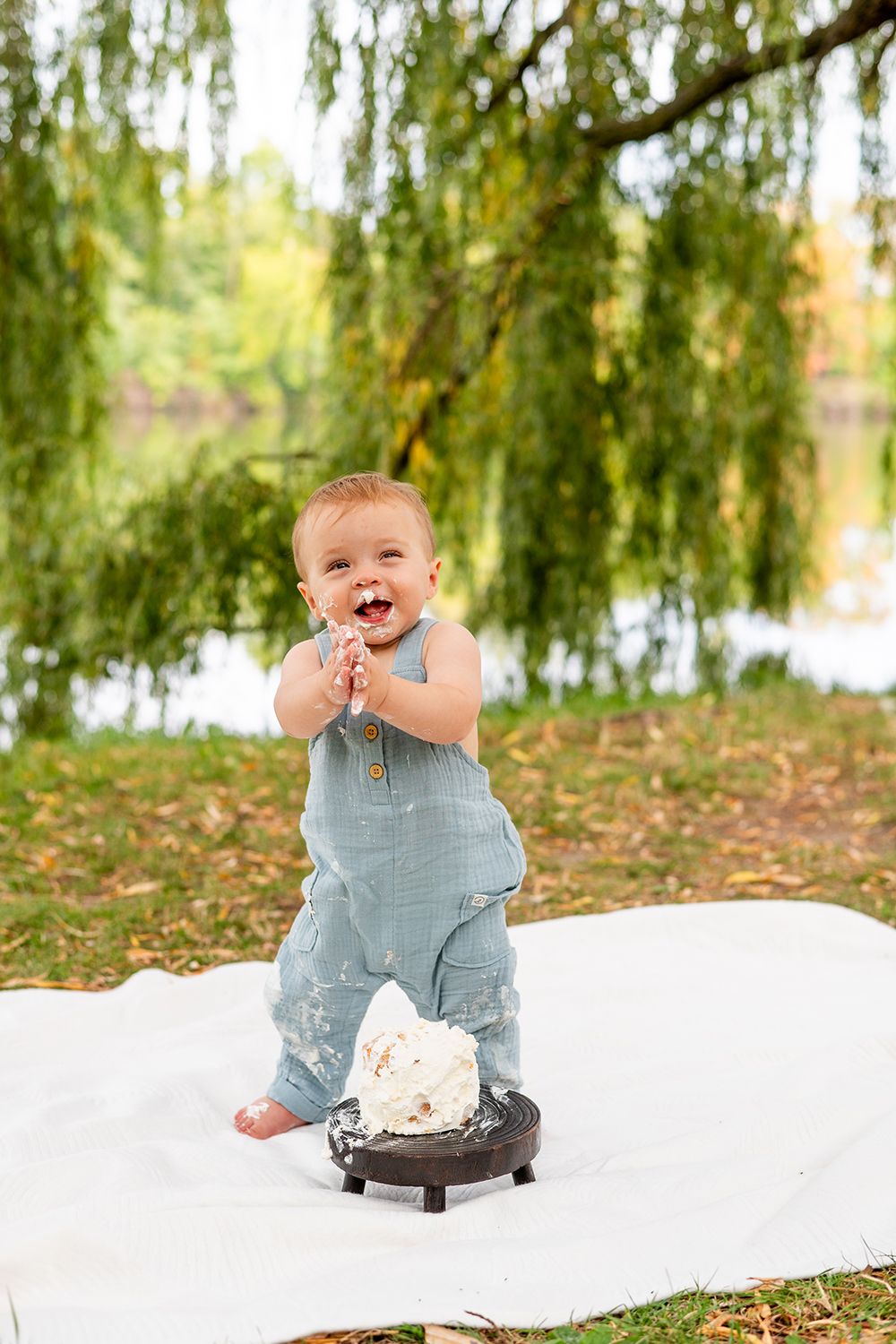 A baby in blue overalls sits on a white blanket with willow trees and grass in the background, smiling with joy.