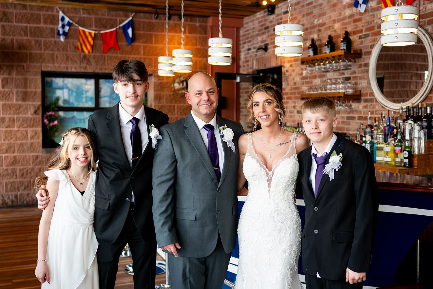 Wedding party members gathered at a rustic brick-walled venue with pendant lighting and bar area in the background.