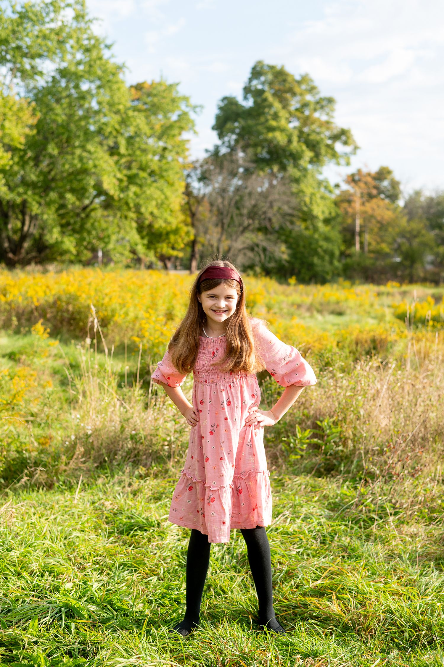 A smiling girl in a pink dress and black tights stands in a sunny field with yellow wildflowers and green trees.