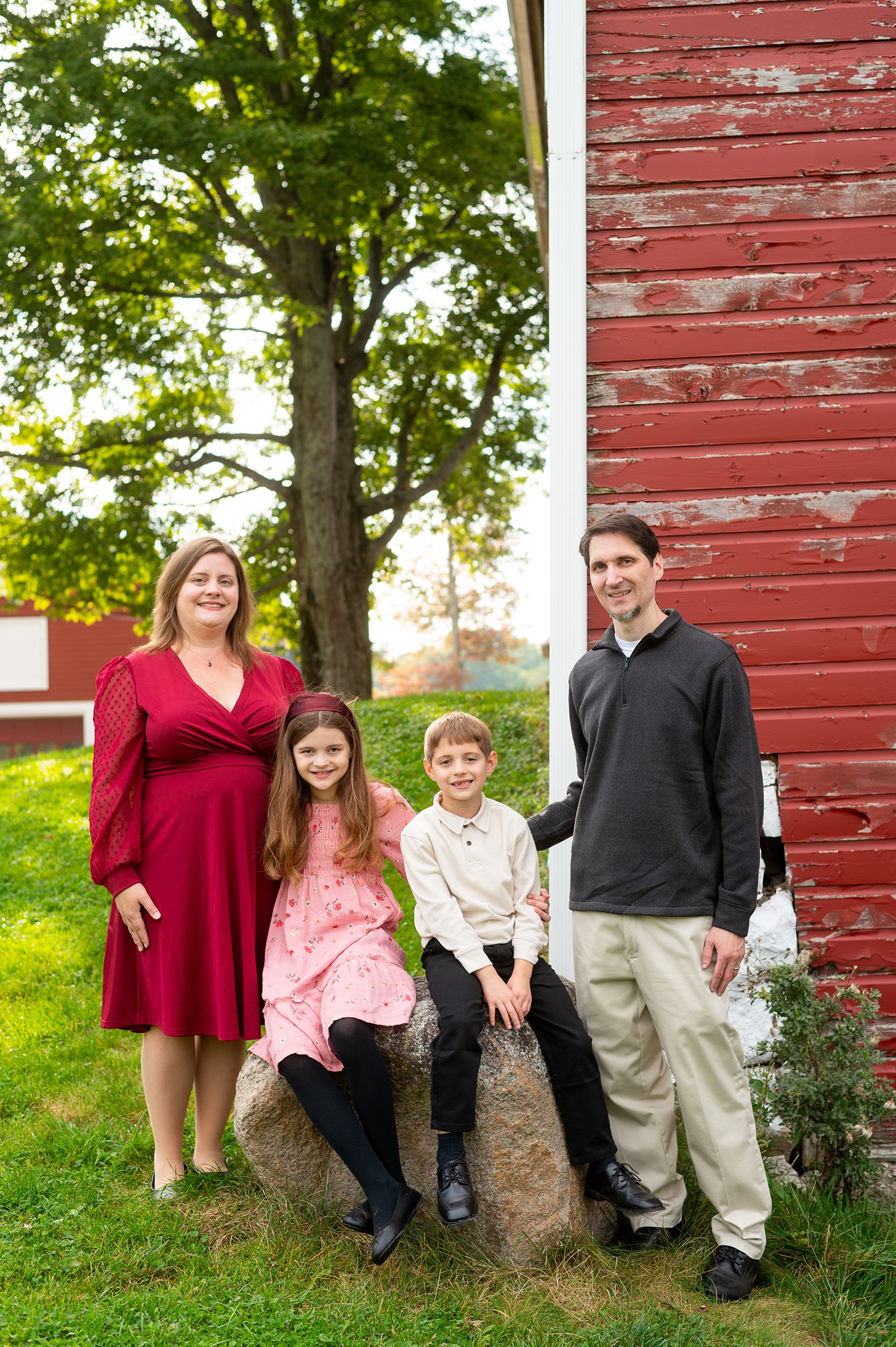 Family of four posing outdoors near a red barn, parents in red dress and dark sweater with two children on a rock.