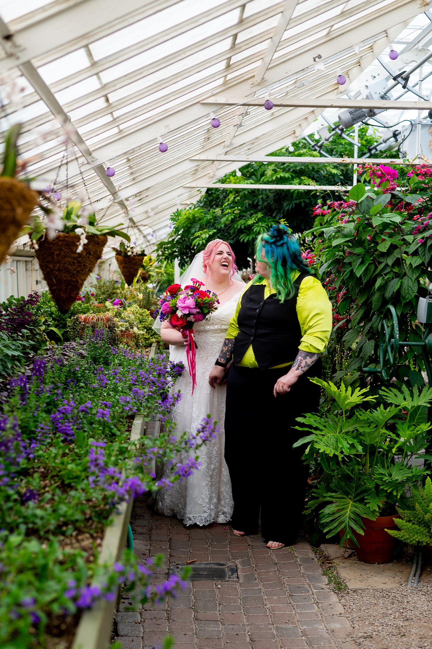 Two brides laugh joyfully in a lush greenhouse, one in a white lace gown with bouquet, the other in yellow jacket with teal hair.