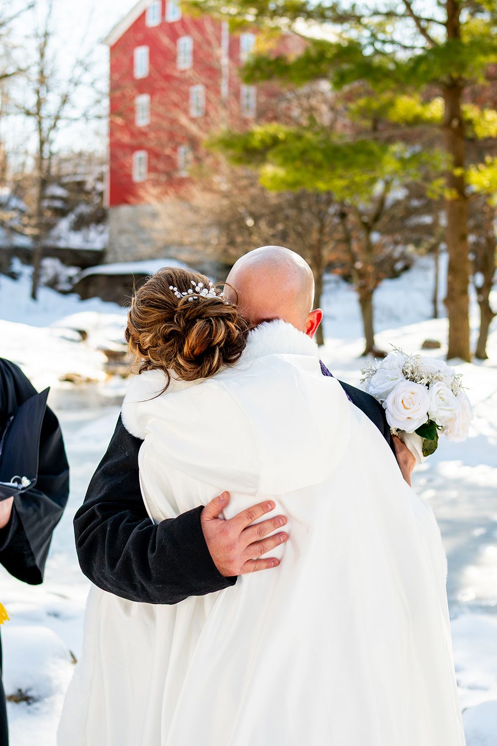 A couple in winter wedding attire dance together in a snowy outdoor setting with a red brick building in the background.