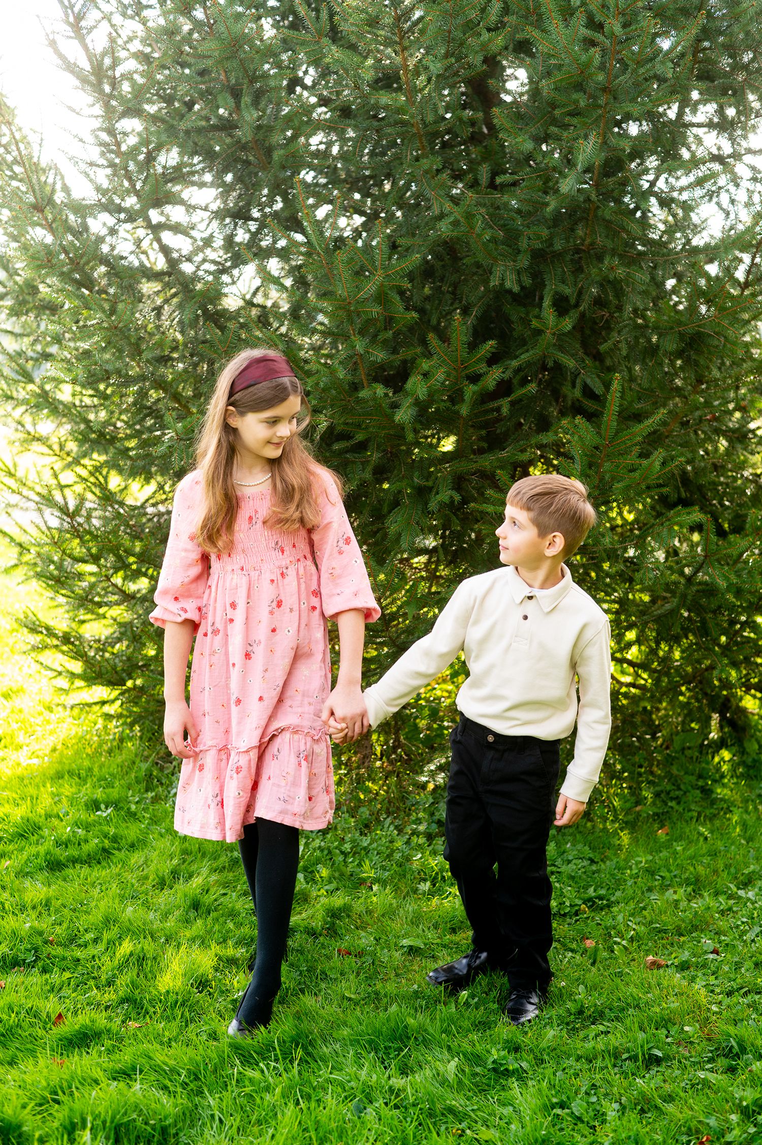 A girl in a pink dress and boy in white shirt hold hands while standing on green grass near evergreen trees.