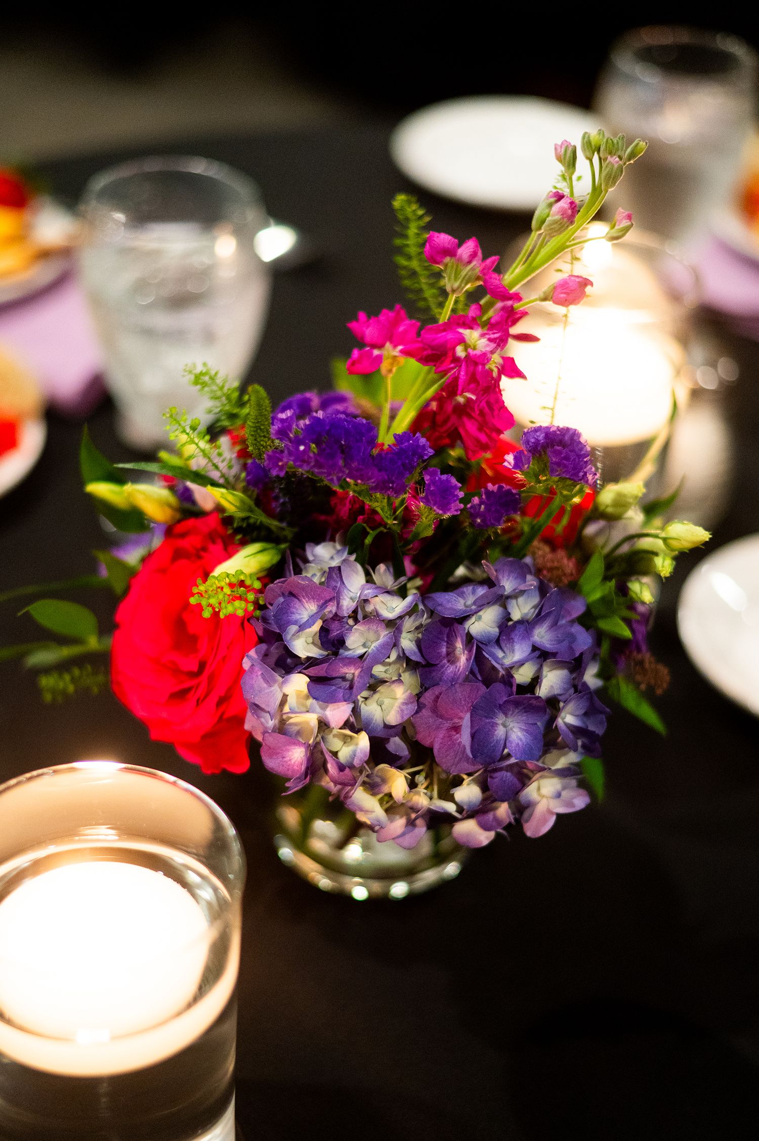 Vibrant floral centerpiece with purple hydrangeas, red roses, and pink blooms surrounded by glowing candles on a dark table.