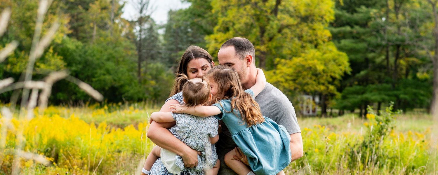 A happy family of four embraces outdoors in a lush green meadow with yellow wildflowers and trees in the background.