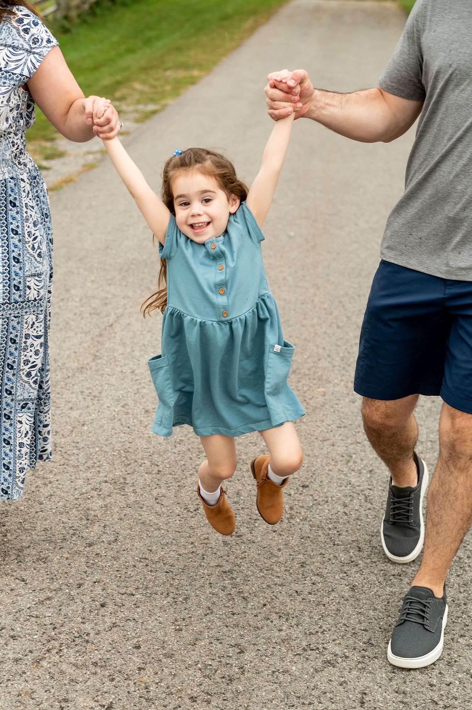 A joyful little girl in a teal dress and brown boots being swung by her parents on a path outdoors.