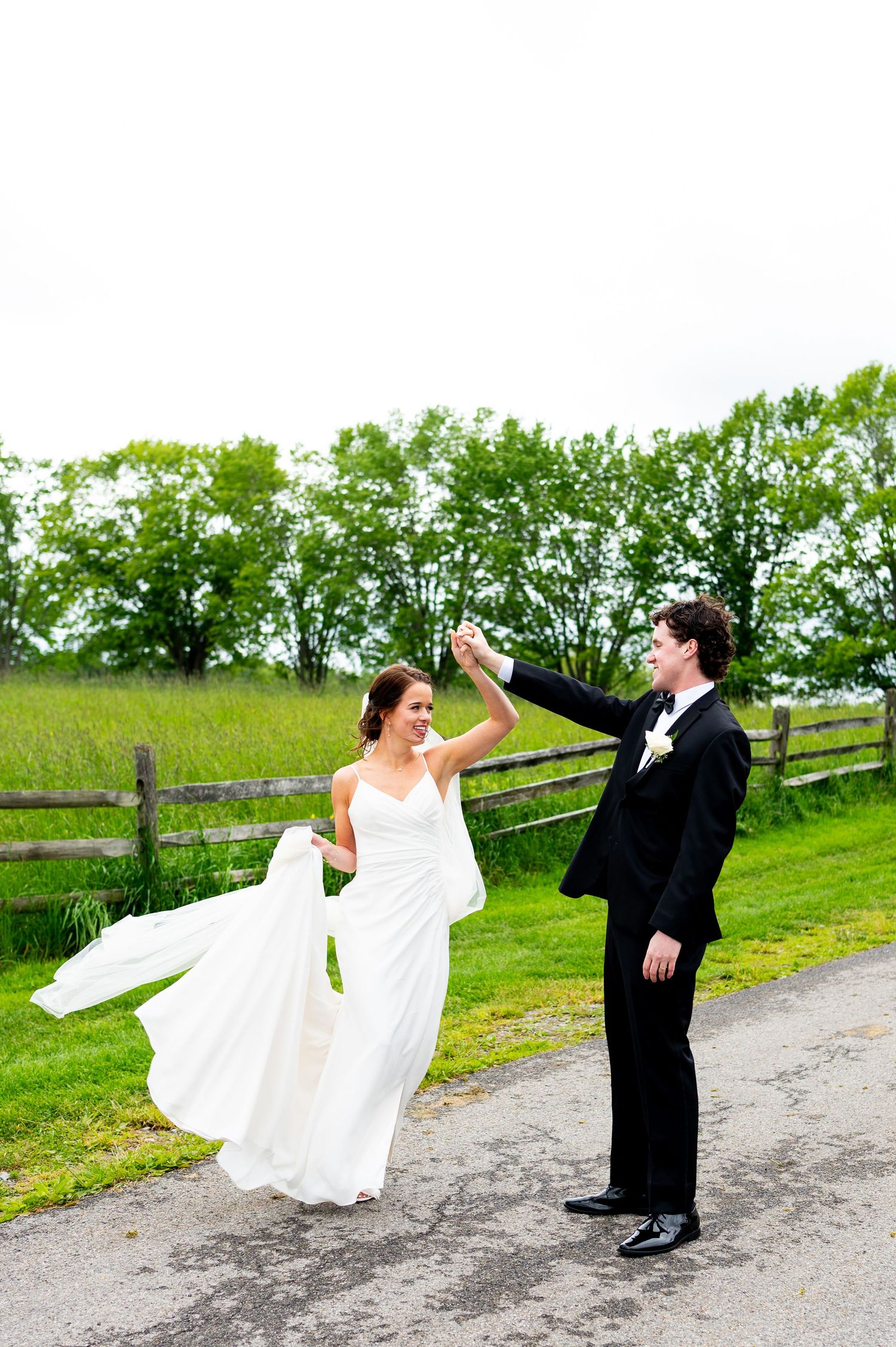 A couple in wedding attire dances playfully on a dirt path surrounded by green fields and wooden fencing.