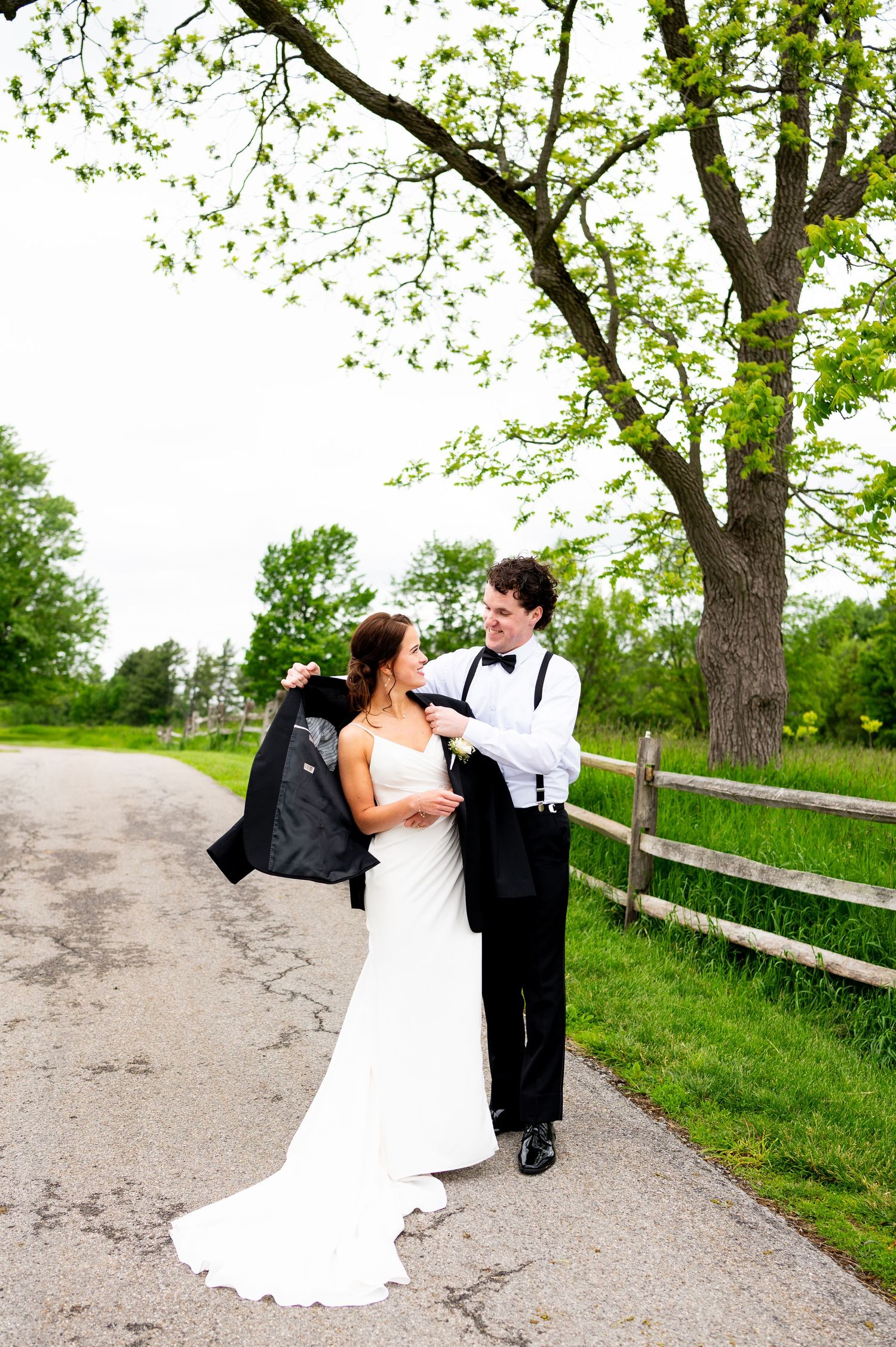 A bride and groom dance playfully on a rustic path bordered by wooden fencing and spring foliage.