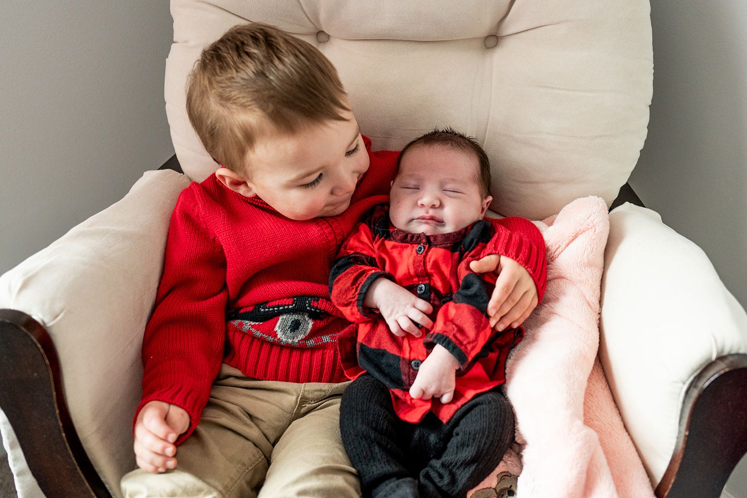 Two young children sit together in a cream colored armchair, wearing matching red outfits.