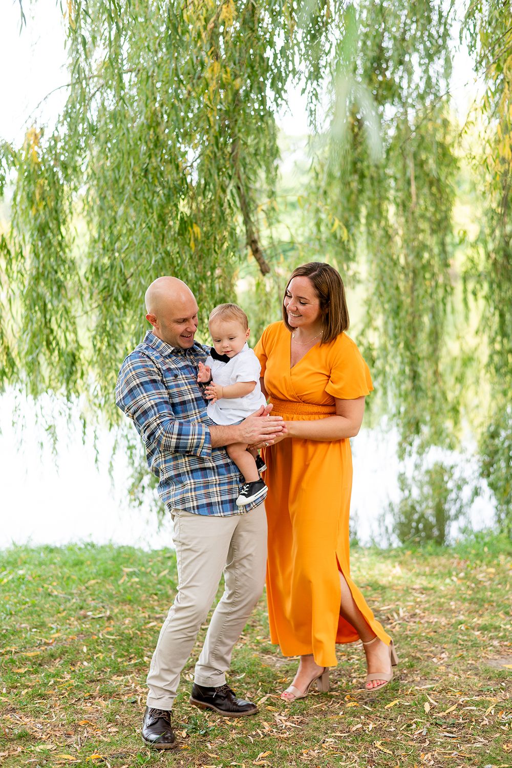 A family in casual attire shares sweet moments together under a willow tree beside a lake.