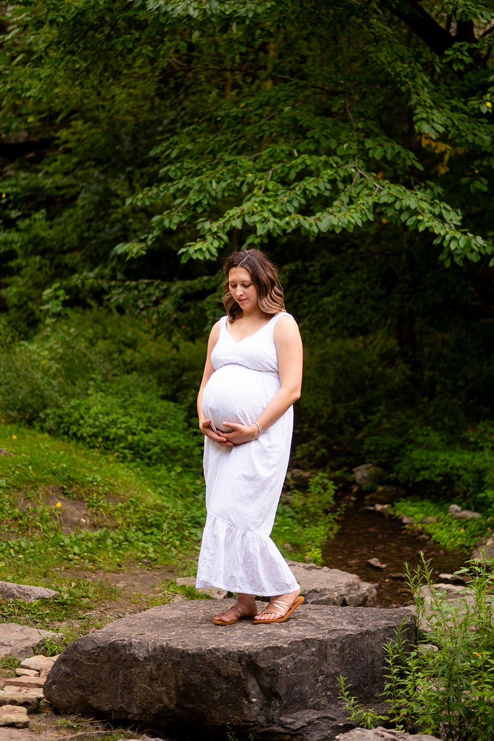 A person in a white summer dress stands on a large rock by a stream in a lush green forest setting.