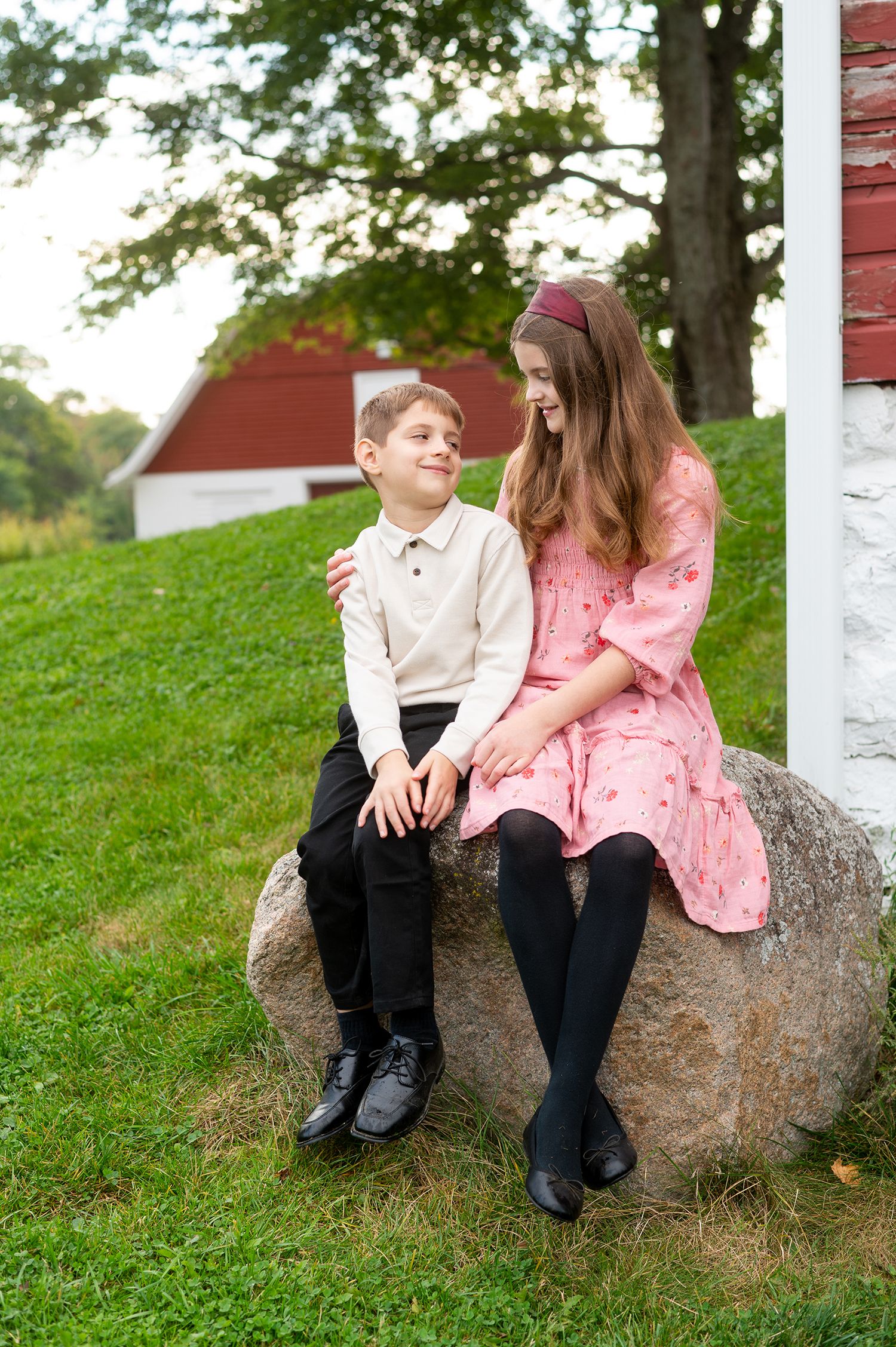 A boy and girl in formal attire sit together on a large rock outdoors near a red barn.