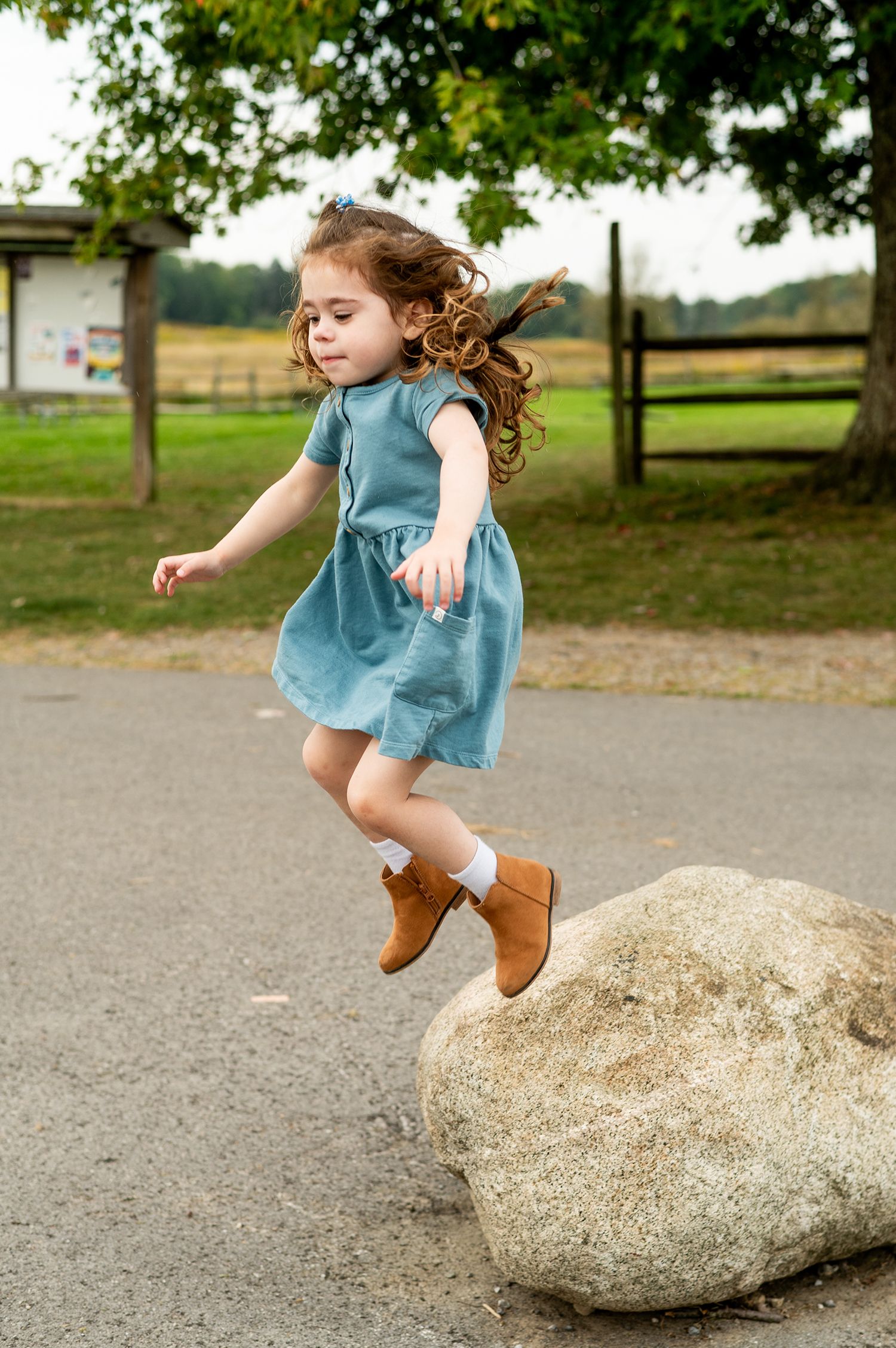 A young girl in a teal dress and brown boots jumping off a large rock in a park setting.