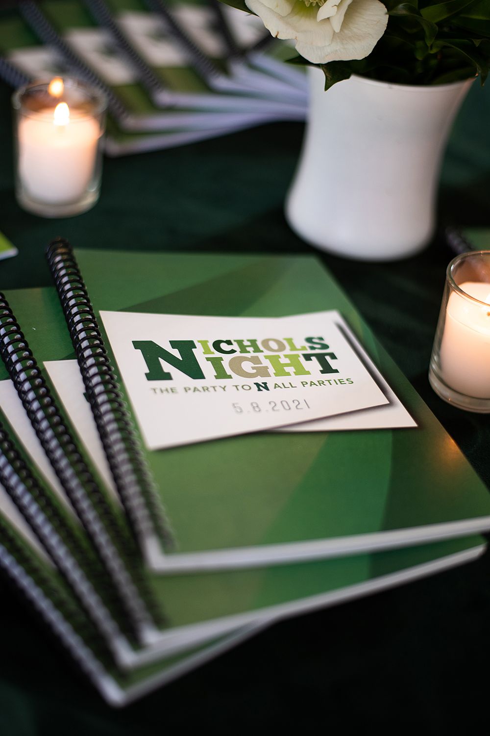 Green notebooks and business material from Nichols Prep displayed on dark table with white candles and floral decor.