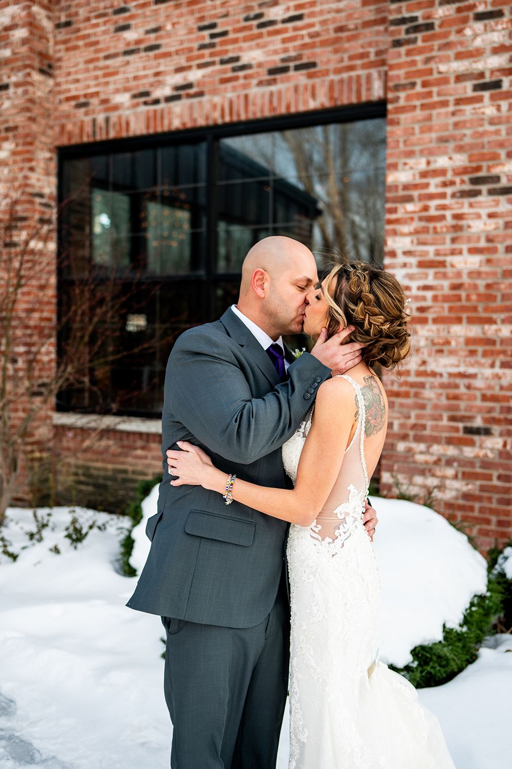 A romantic winter wedding portrait with a couple embracing against a red brick building with snow on the ground.