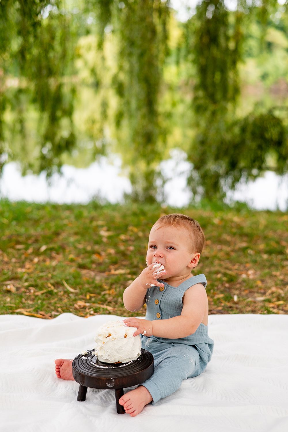Baby in blue romper sitting on white blanket with cake smash setup under willow tree branches.