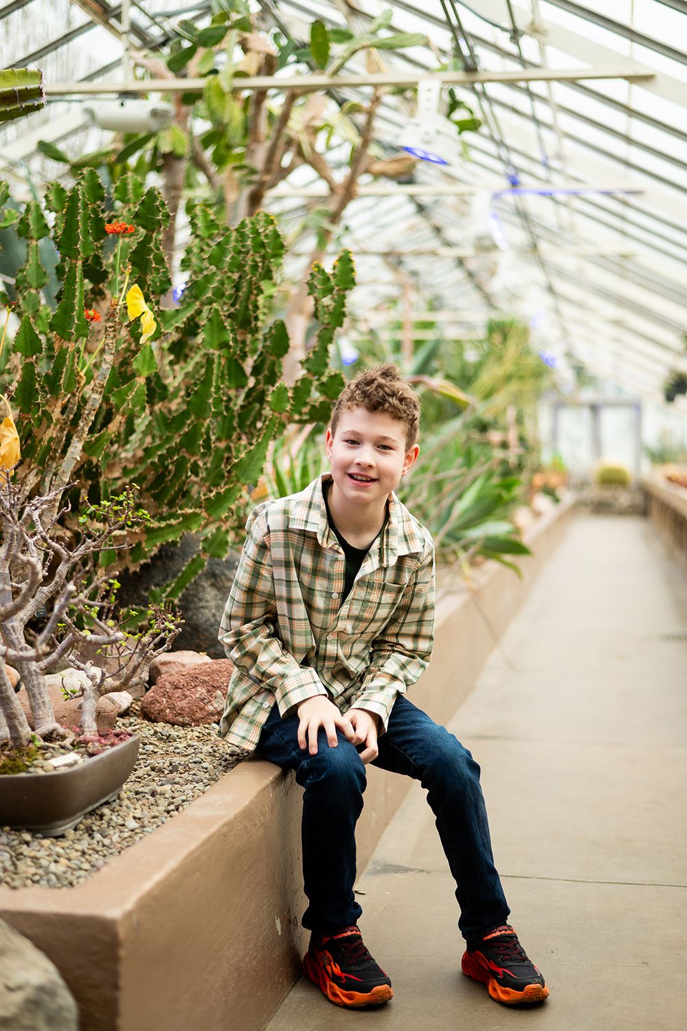 Person sits on concrete ledge inside greenhouse with desert plants and cacti in plaid flannel shirt and orange shoes.