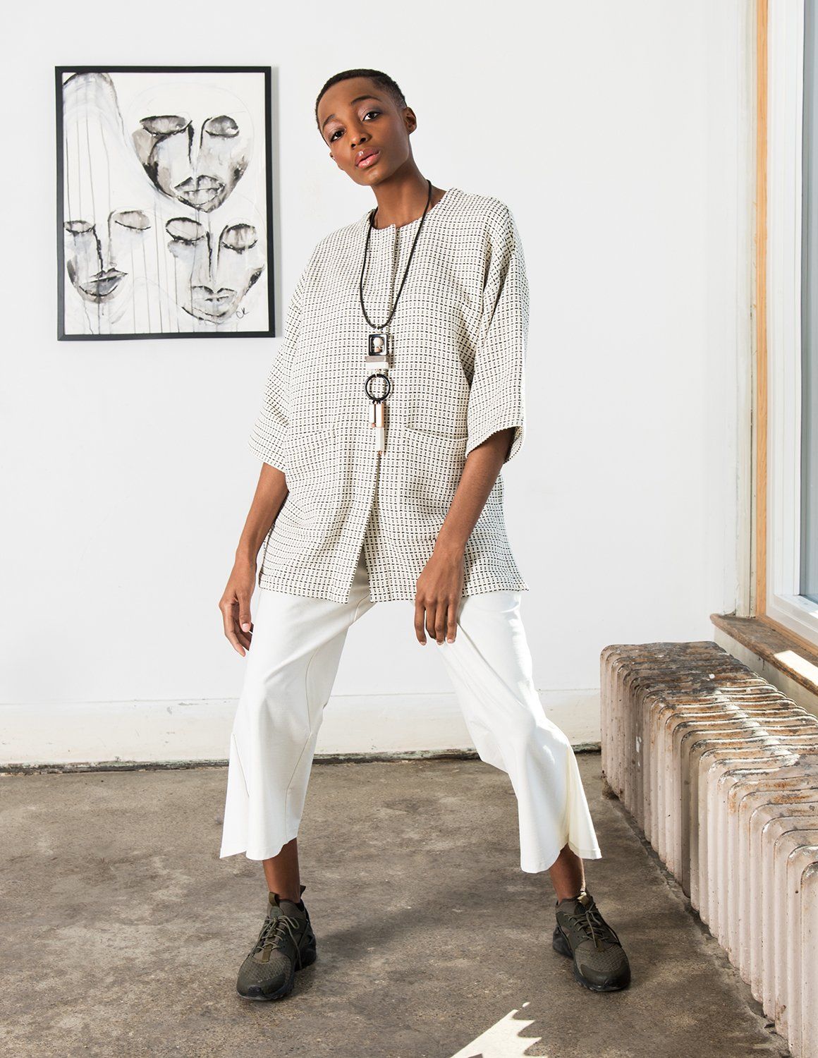 A person in a white textured tunic and wide-leg pants poses casually next to abstract face artwork and a radiator.