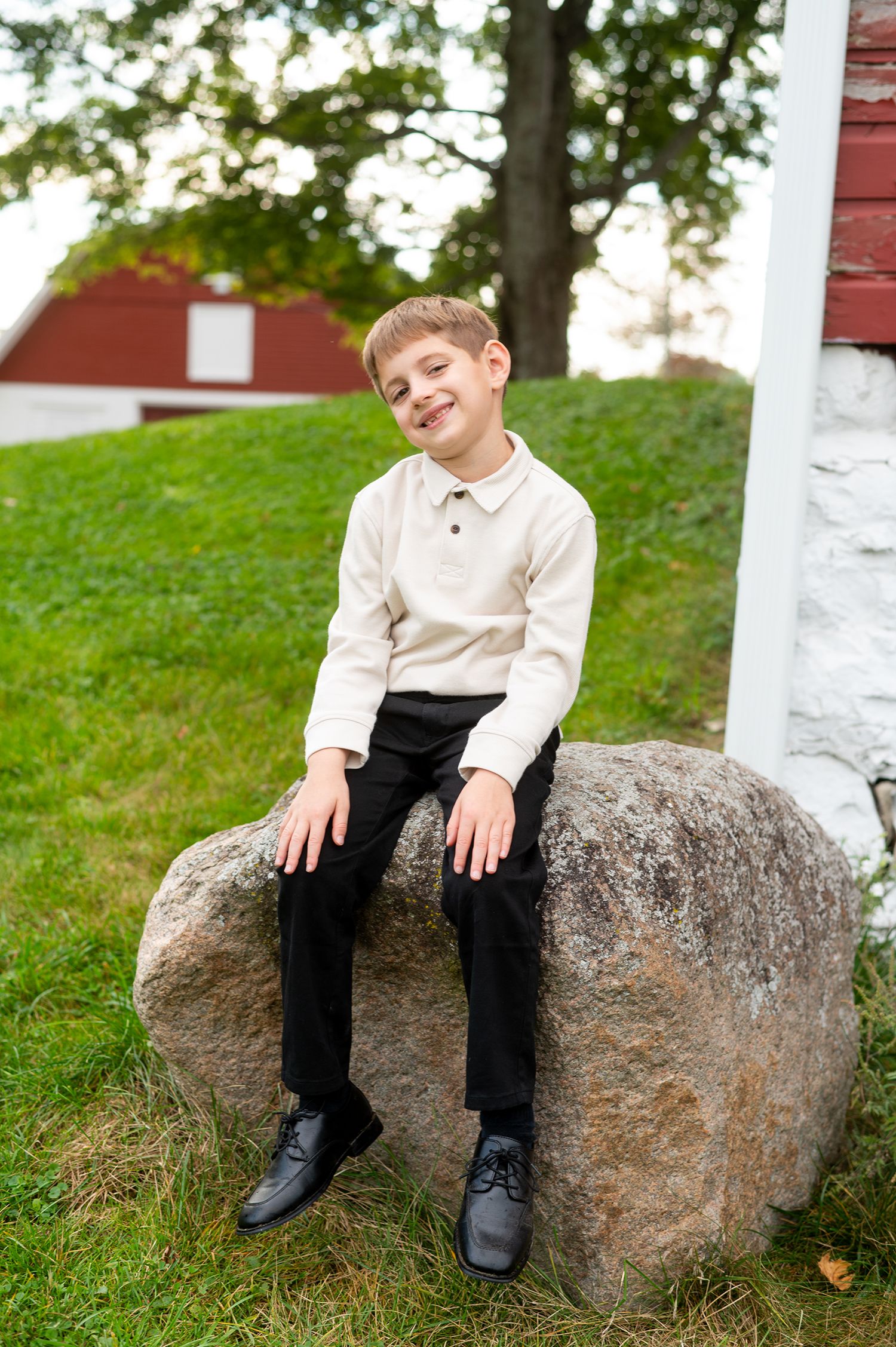 A smiling boy in a cream sweater and black pants sits on a large rock near a red barn.