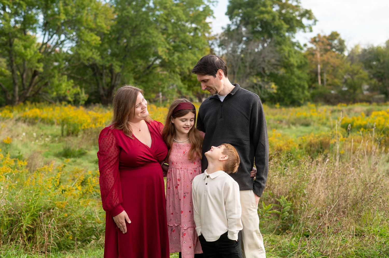 Family of four posing outdoors in a green meadow, mother in red maternity dress with children and tall father.