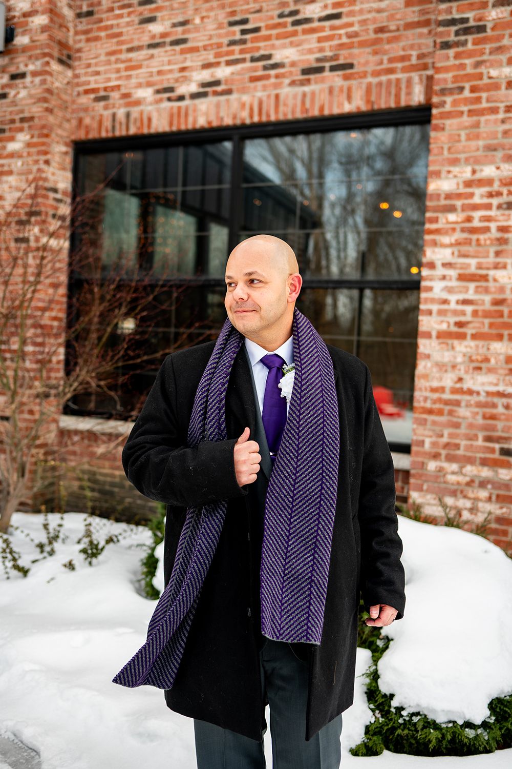 Professional winter portrait of a person wearing a black wool coat and purple striped scarf against a snowy brick backdrop.