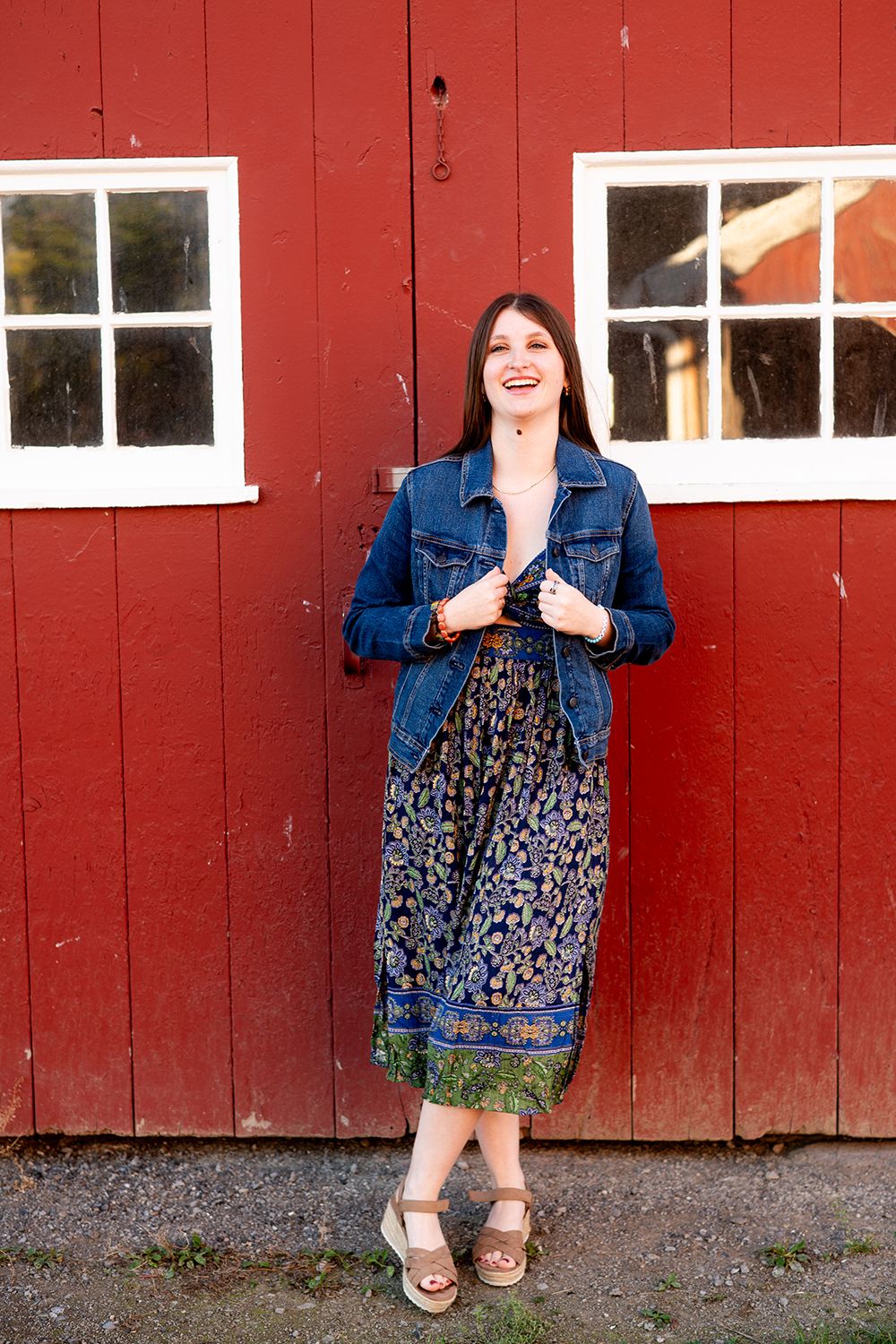 Person wearing a denim jacket and floral dress poses in front of a red barn wall with white windows.