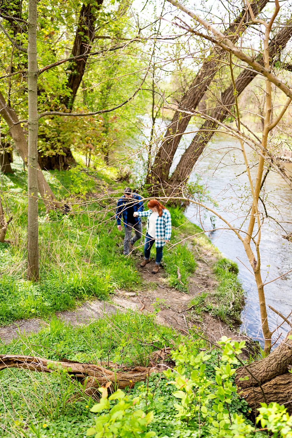 Two hikers walk along a narrow dirt path beside a river on a spring day surrounded by green foliage and trees.
