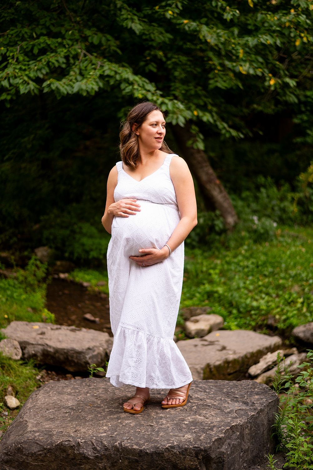 A pregnant woman in a white flowing sundress stands on a large rock in nature, cradling her belly in a serene setting.