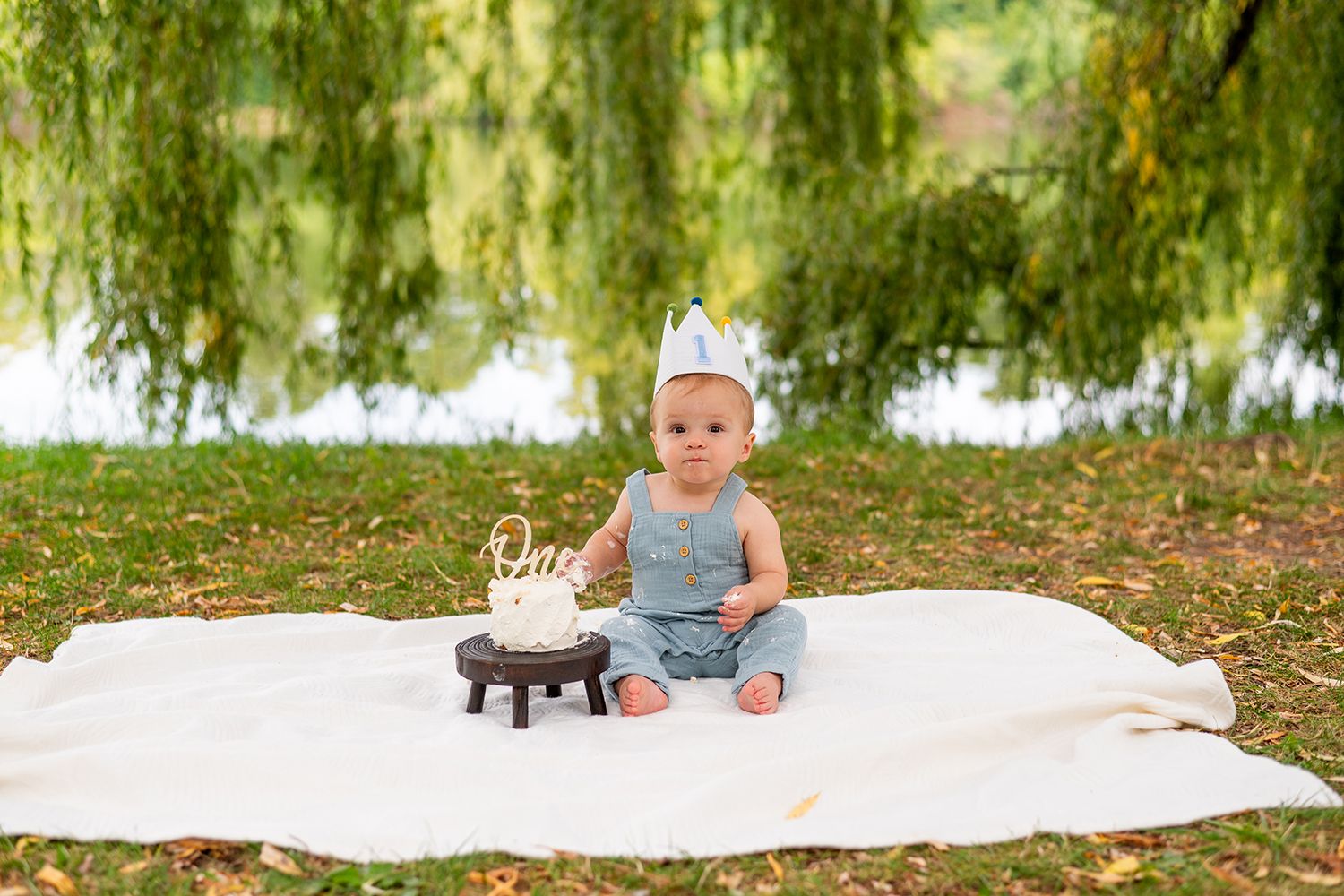 A toddler in denim overalls and grey shirt sits on a white blanket in a park setting outdoors.