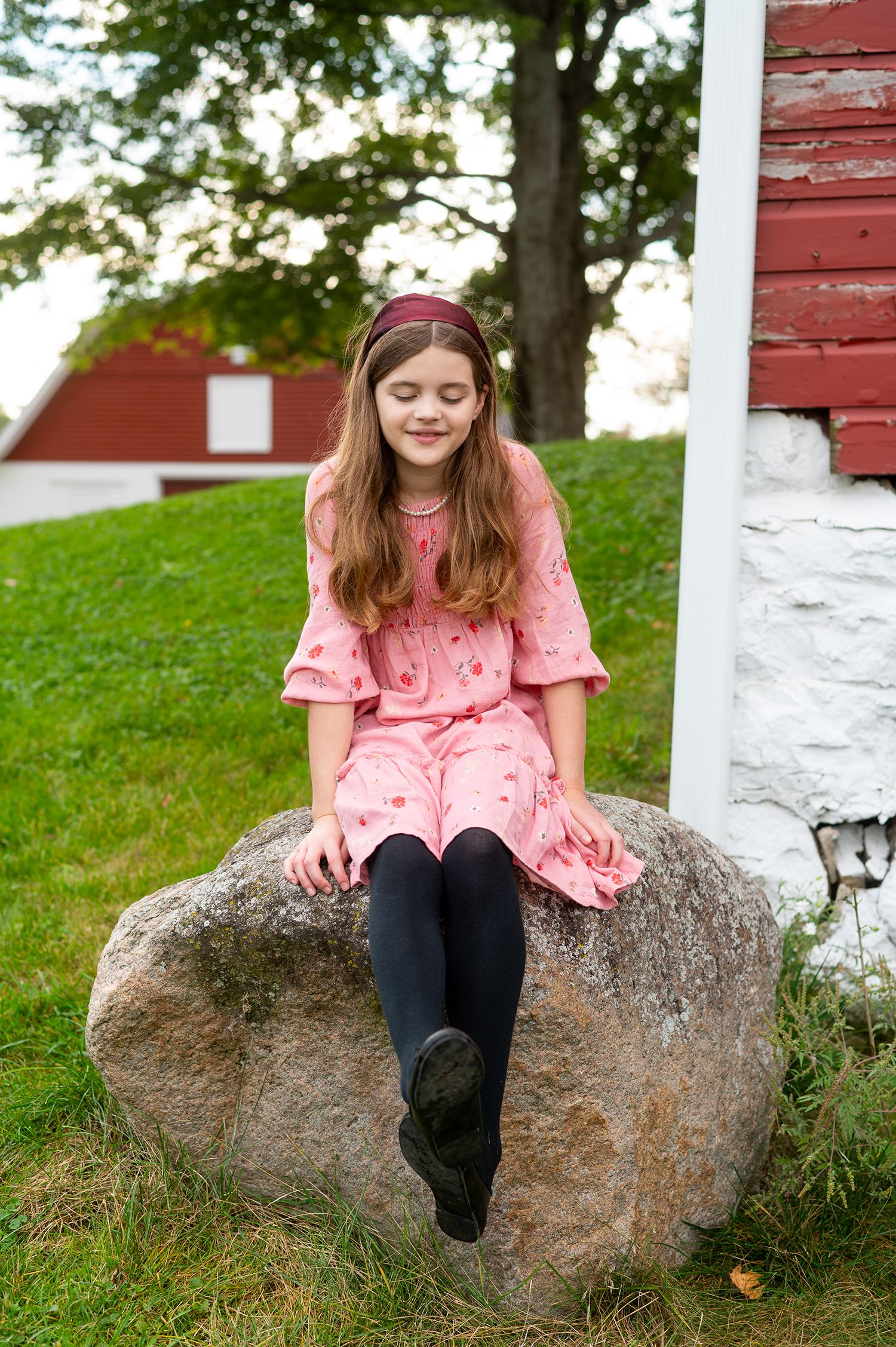 Young girl in pink dress and black tights sitting on a rock near a red barn with eyes closed.