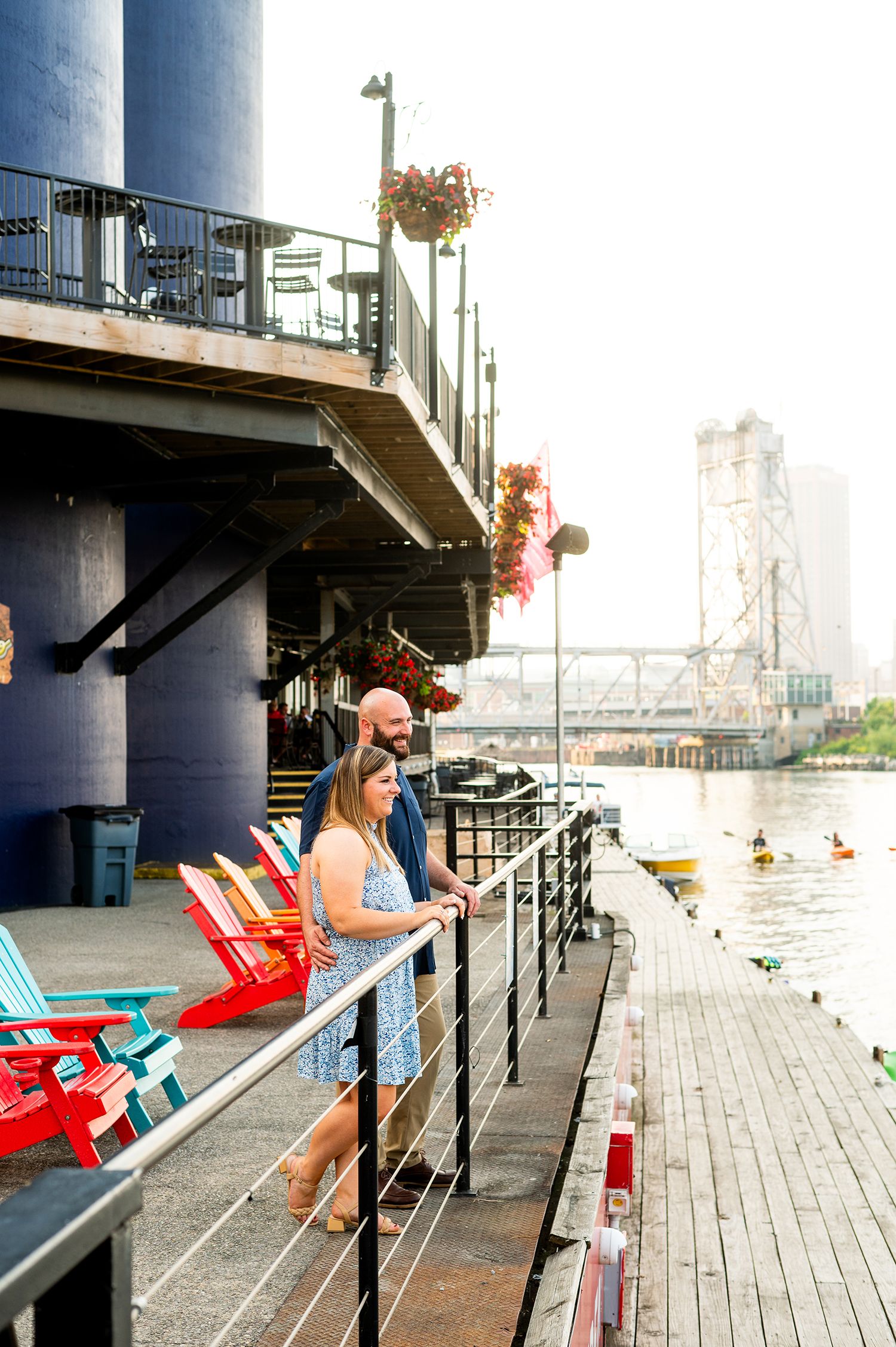 A couple stands on a waterfront deck with colorful Adirondack chairs, overlooking a river with a bridge in the misty background.