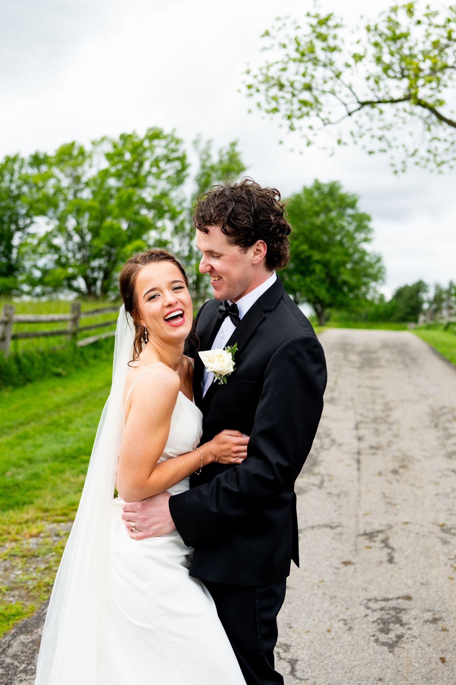 A joyful wedding couple shares candid laughter together on a country path surrounded by green spring foliage.
