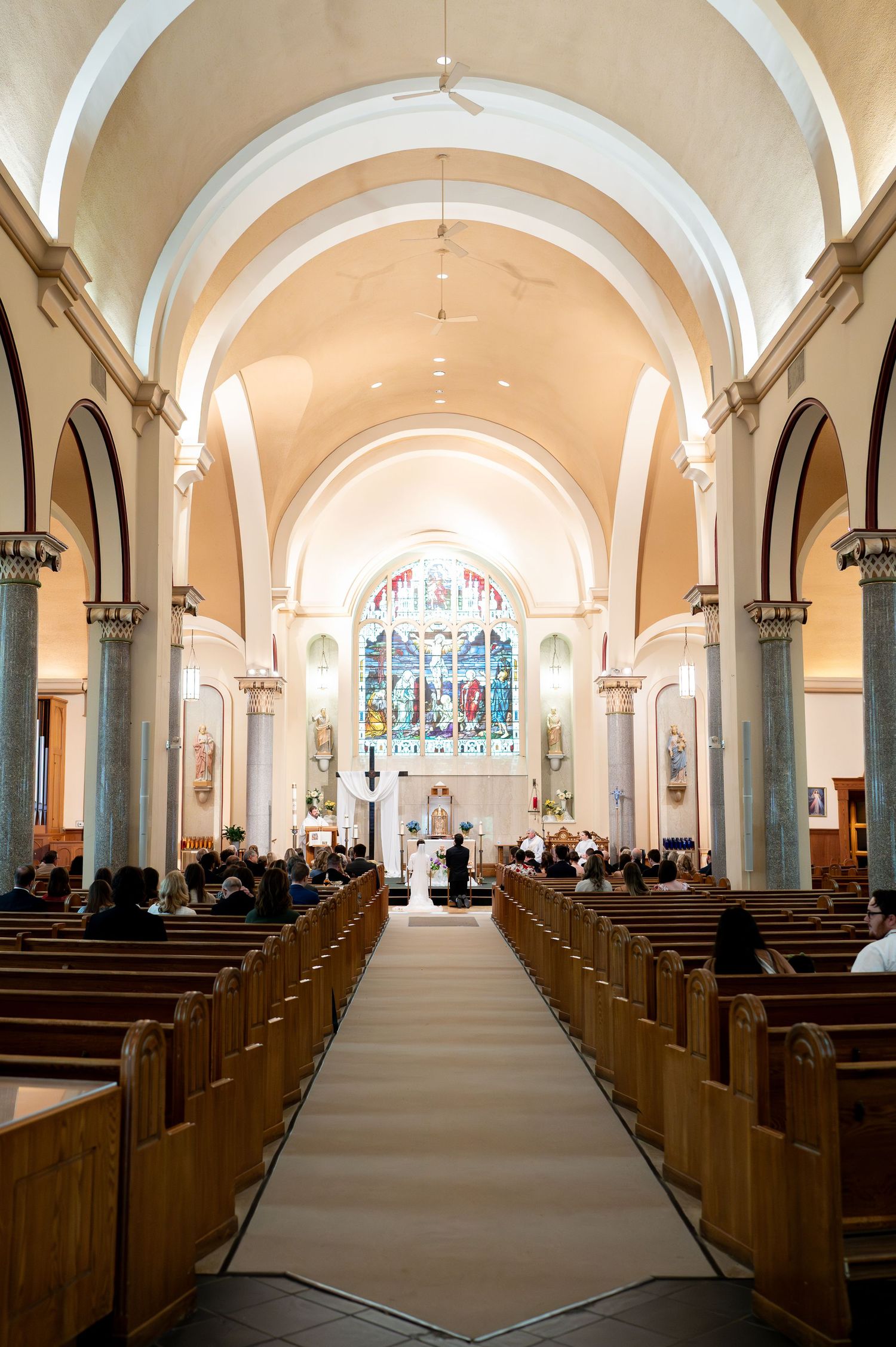 Interior view of a church ceremony with vaulted ceilings, wooden pews, and a beautiful stained glass window at the altar.