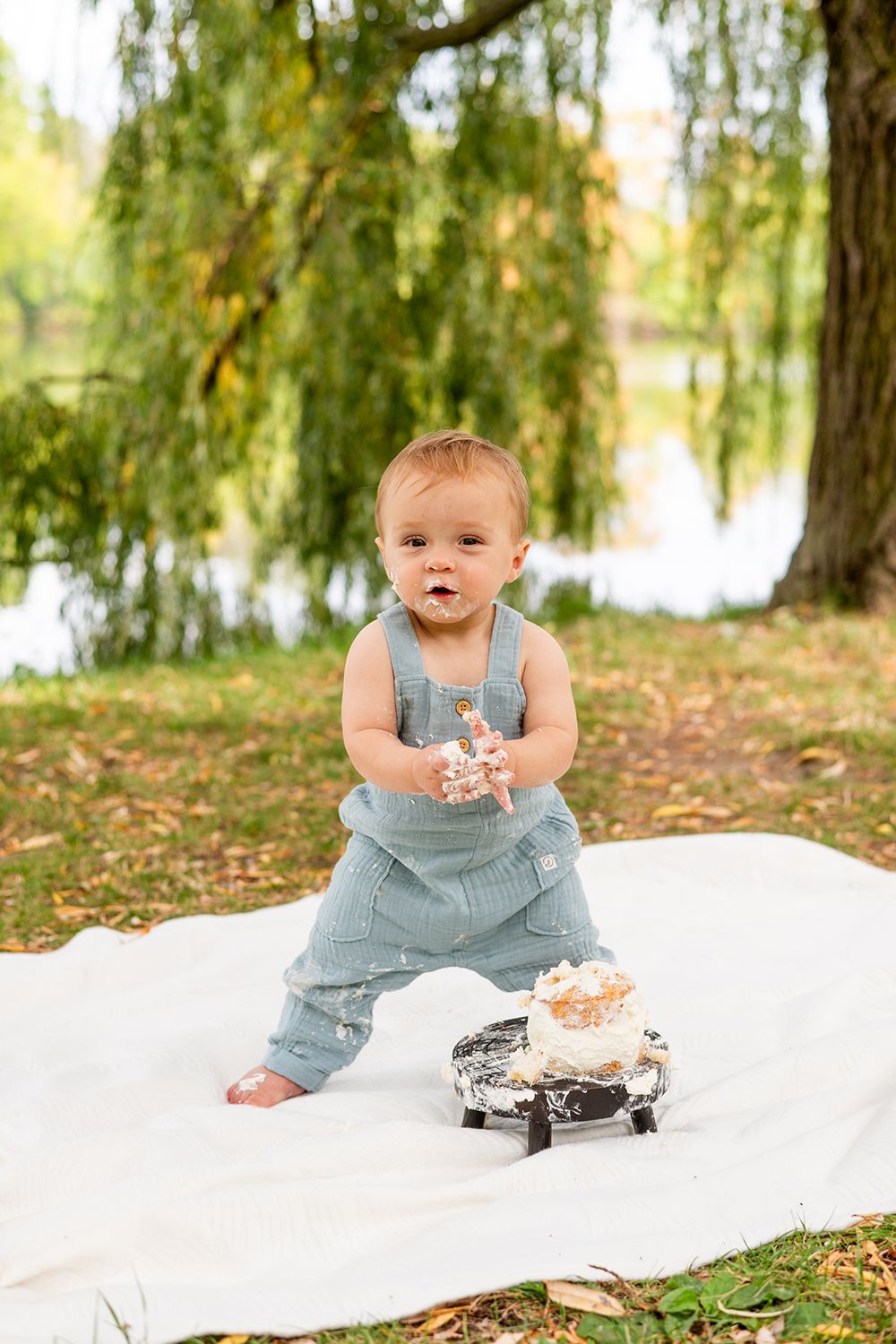 A toddler in blue denim overalls stands barefoot on a white blanket next to a smash cake outdoors.