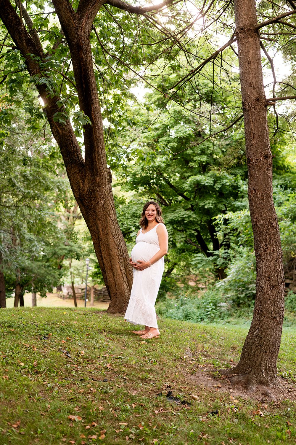 A pregnant woman in a white dress stands between two large trees in a lush green park setting during summer.