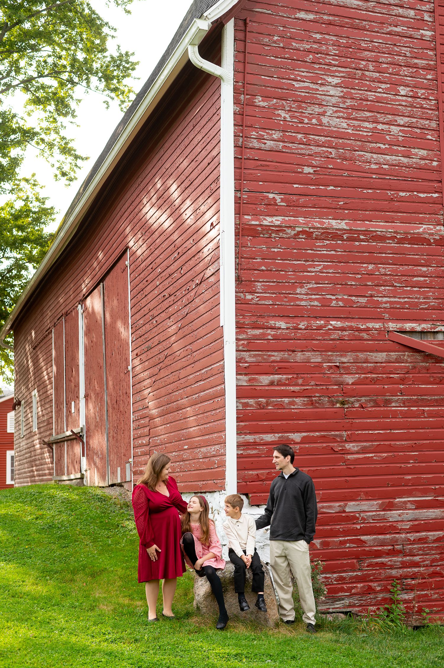 Family of four posing beside a rustic red barn with peeling paint on a green lawn.
