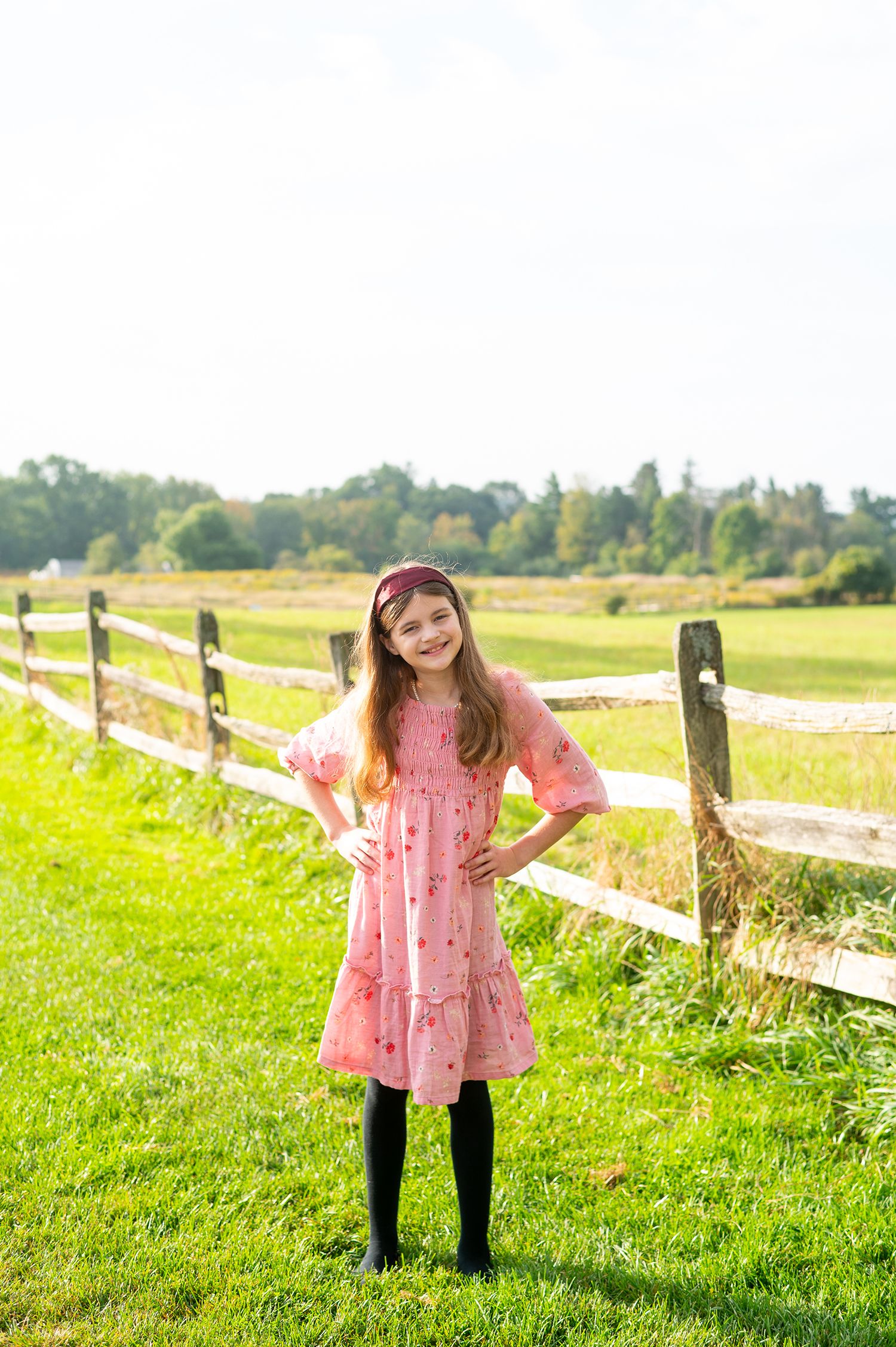 Young girl in pink dress and black tights poses by wooden fence in sunny green field.