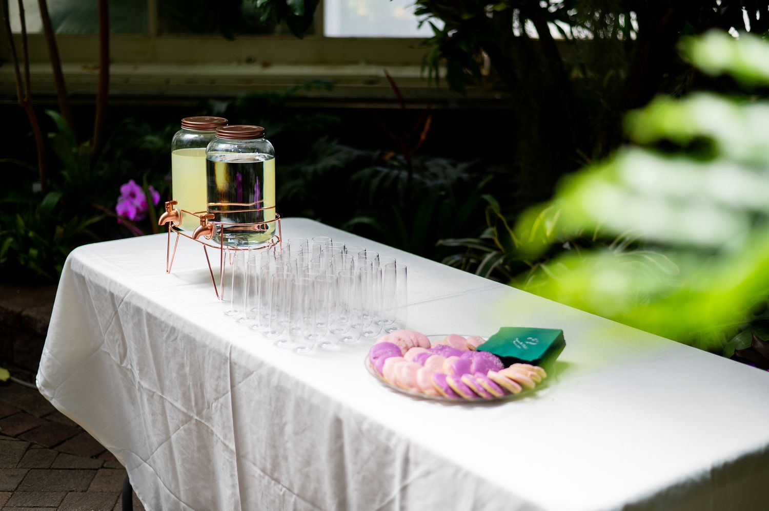 Elegant outdoor event table with white linen, glass drink dispenser, champagne flutes, and floral arrangement.