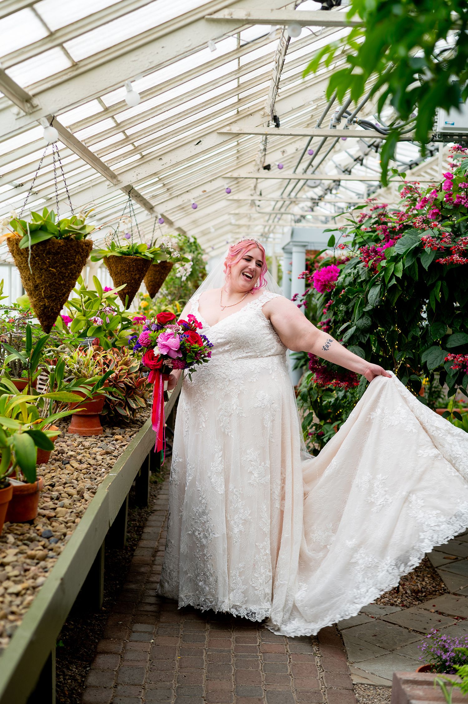 Joyful bride with pink hair in lace wedding gown holding bouquet in a lush greenhouse setting.