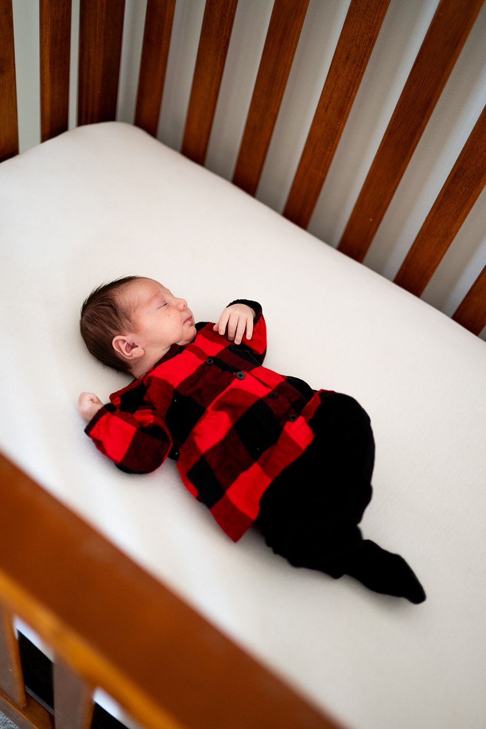 A sleeping baby wearing red and black buffalo plaid lies in a wooden crib with white bedding.