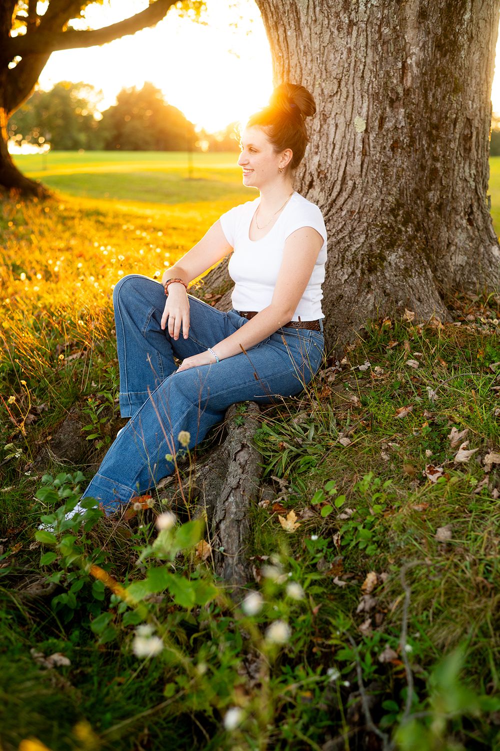 Sitting against a tree trunk at golden hour, someone in jeans relaxes in a grassy field with warm sunlight filtering through.