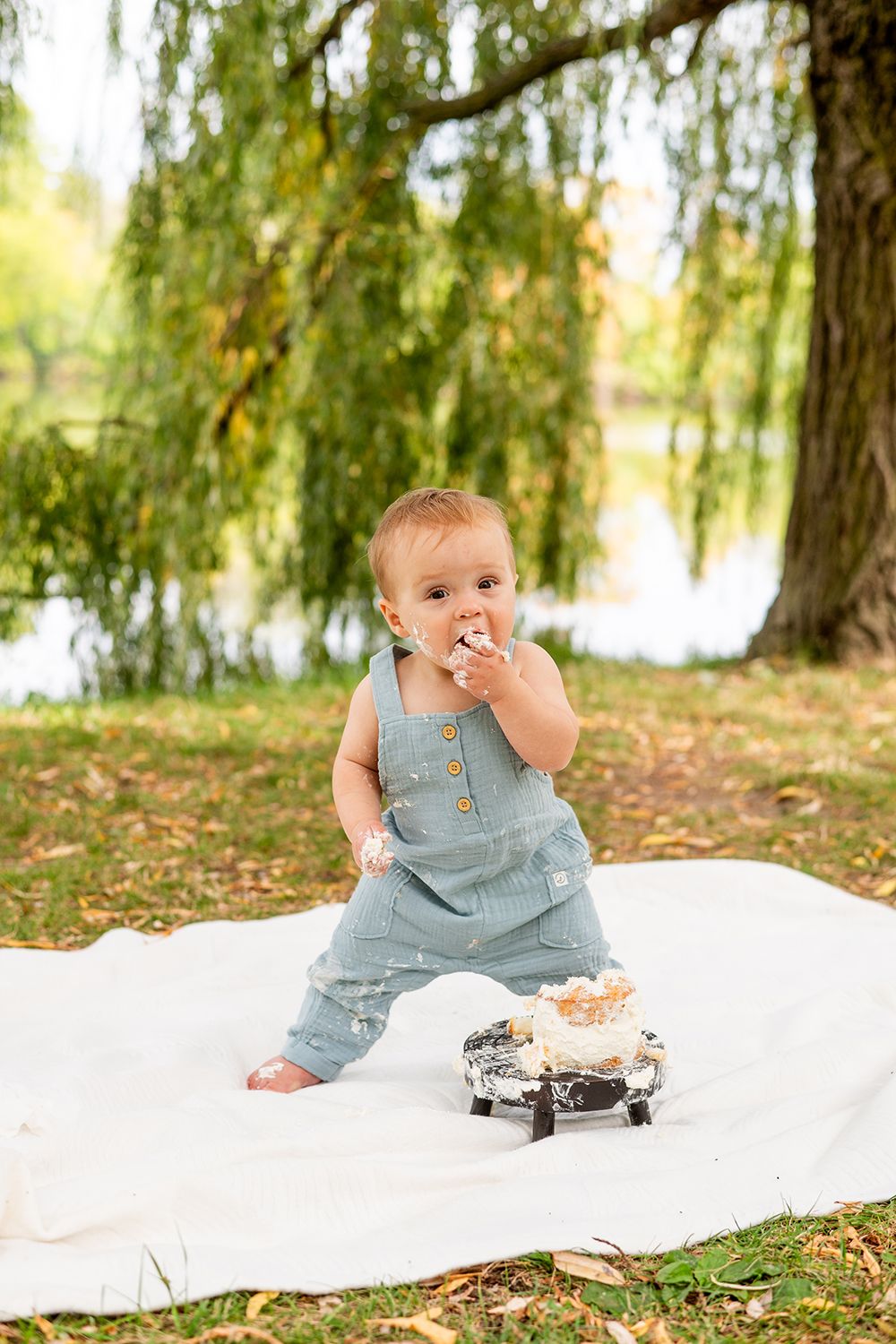 A small child in denim overalls enjoys snacks while standing on a white blanket outdoors near a willow tree.