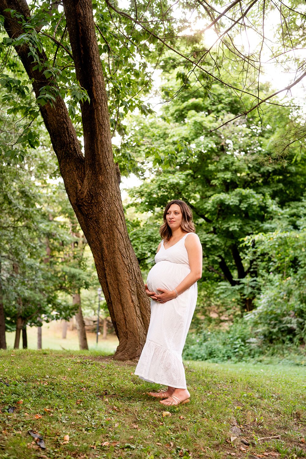 Woman in white dress poses gracefully on grassy hillside surrounded by sunlit trees in a summer setting.