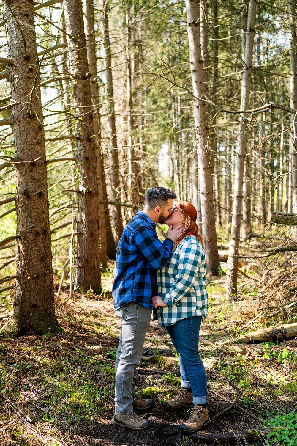 A couple in matching blue plaid flannel shirts sharing an intimate moment in a sunlit pine forest.