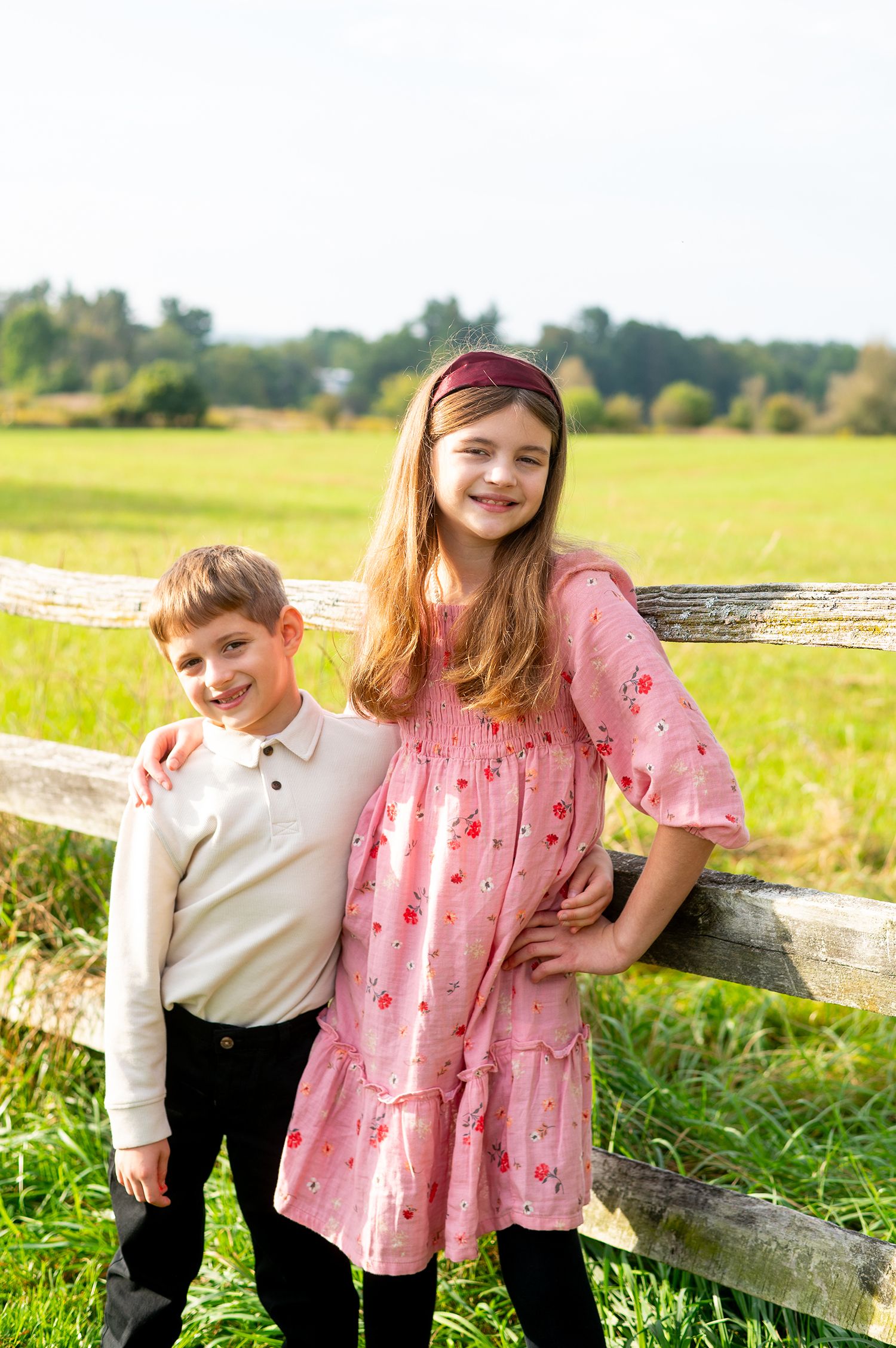 Two smiling children pose by a wooden fence in a sunny green field, girl in pink floral dress, boy in white polo shirt.