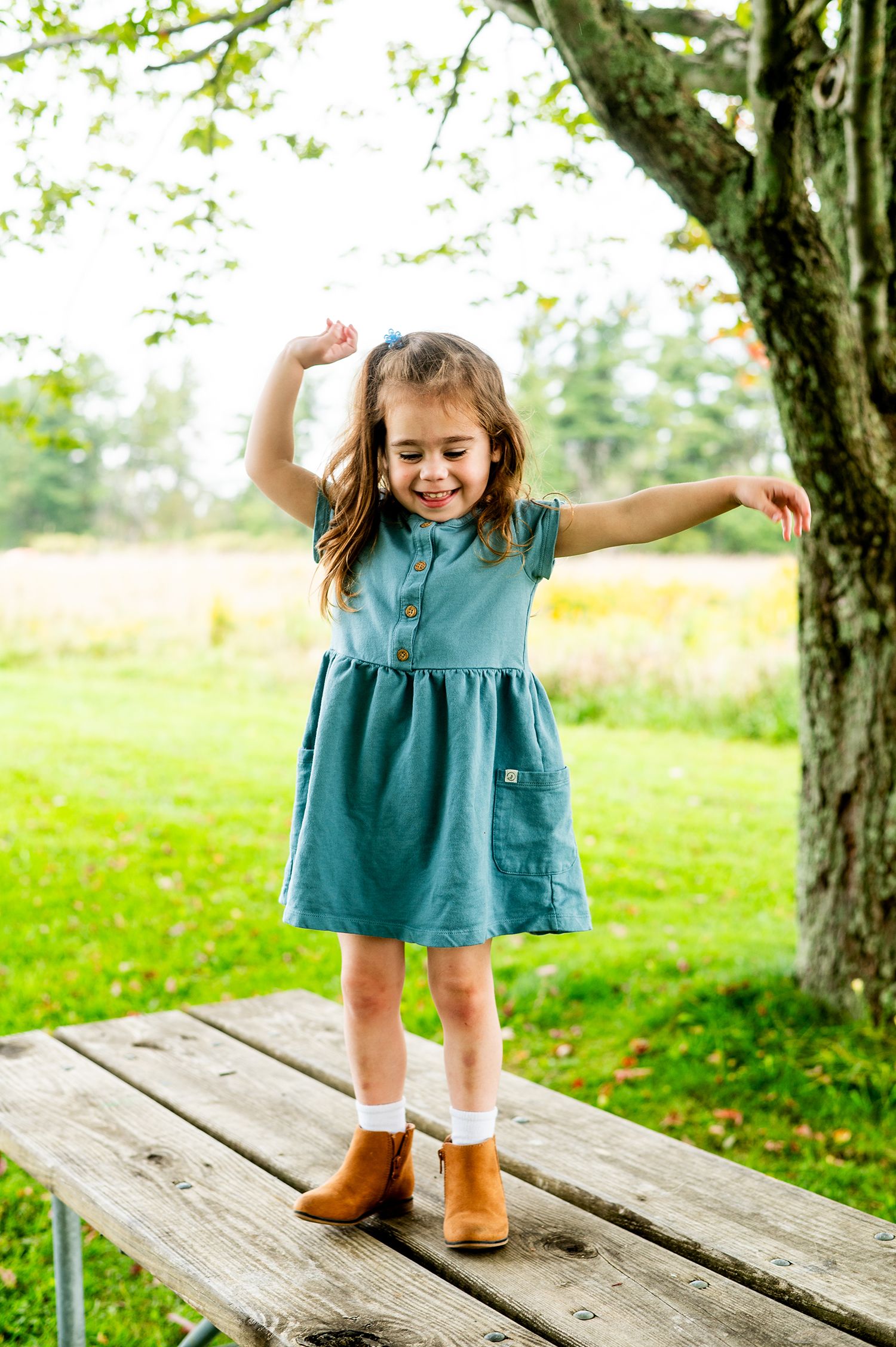 Young girl in teal dress and brown boots dancing on a wooden picnic table outdoors near a tree.