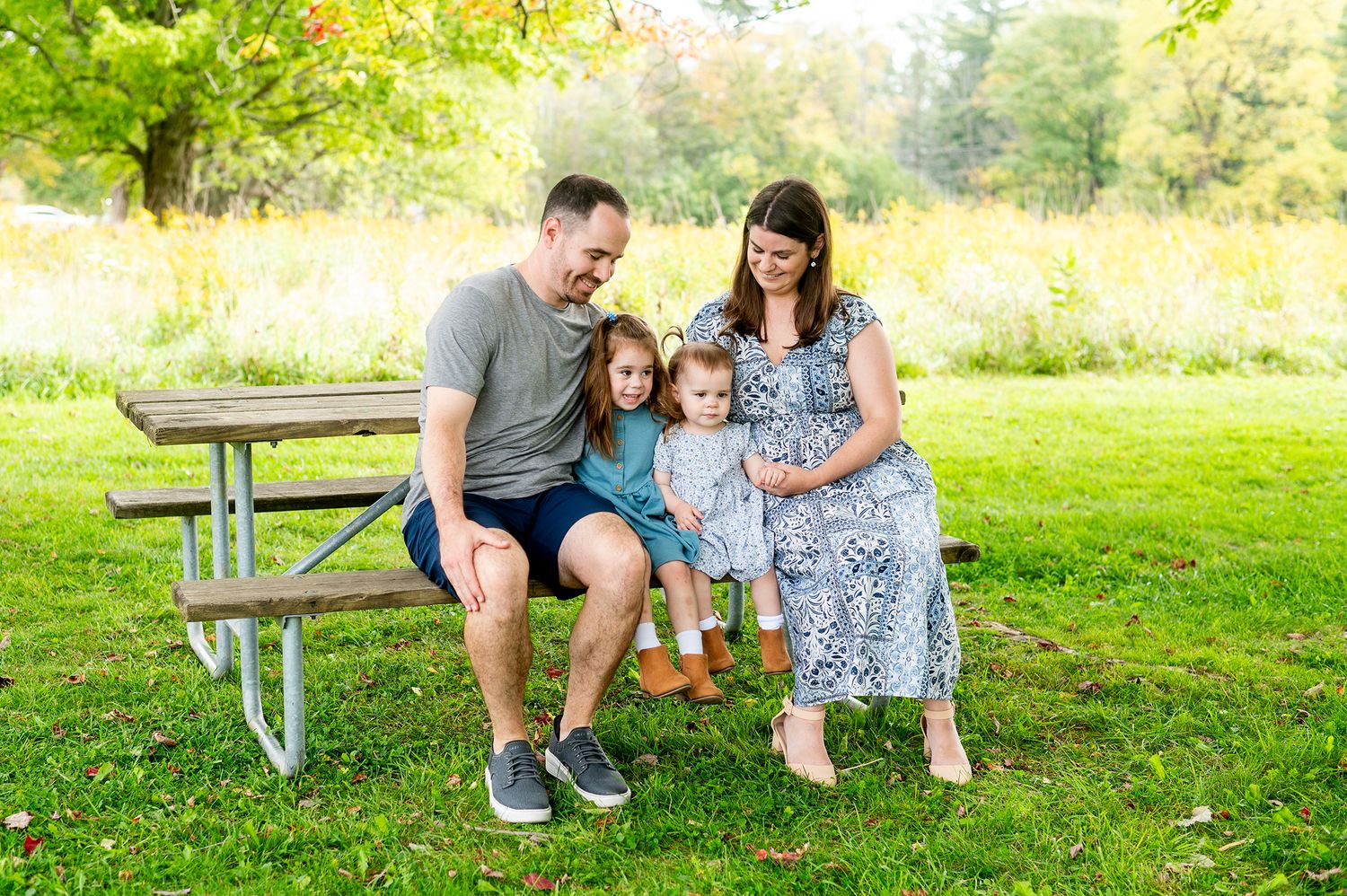 Family of four smiling together on a picnic bench in a lush green park on a sunny day.