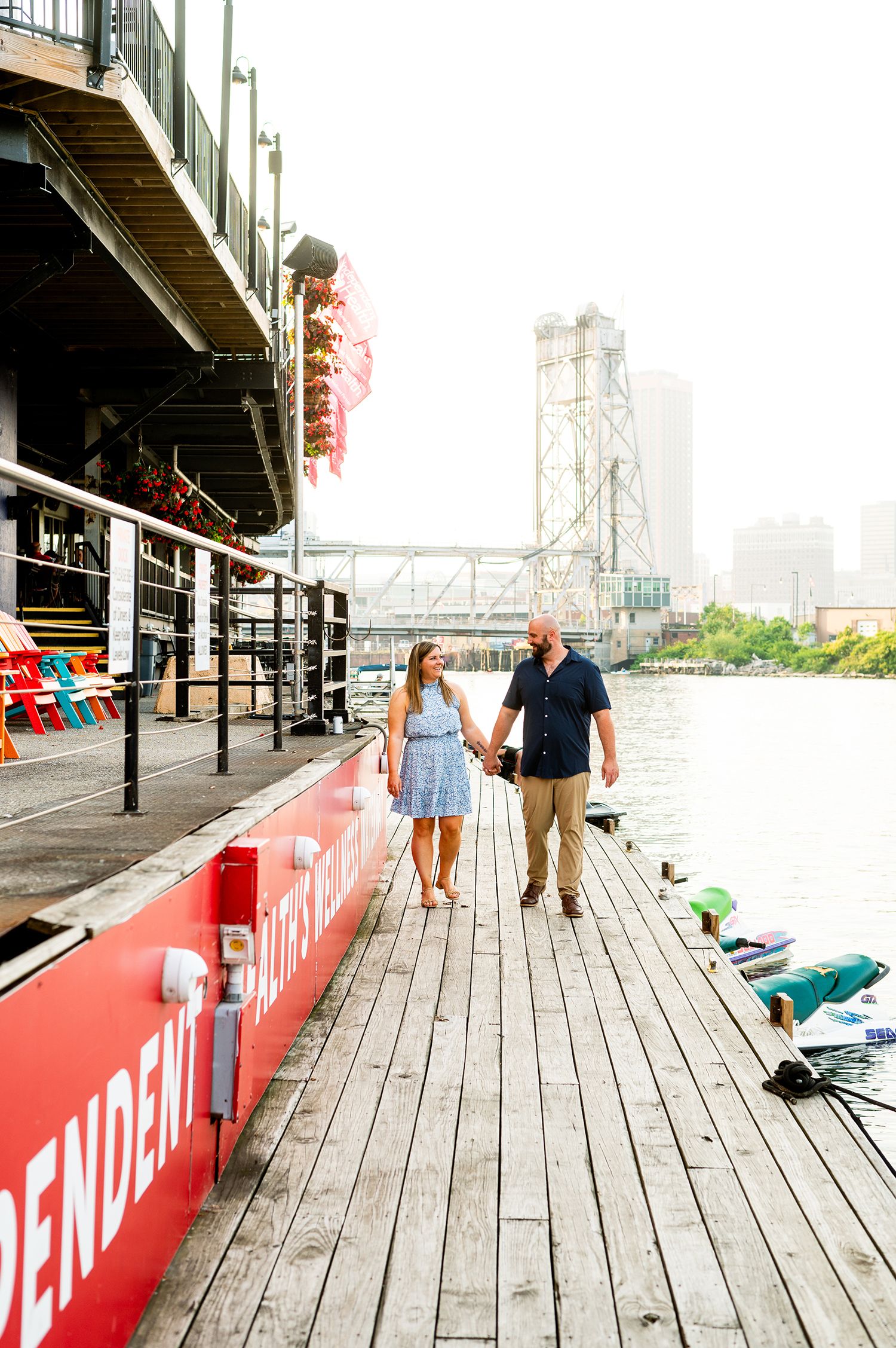 Couple holds hands walking on wooden dock by red riverboat with bridge skyline backdrop.