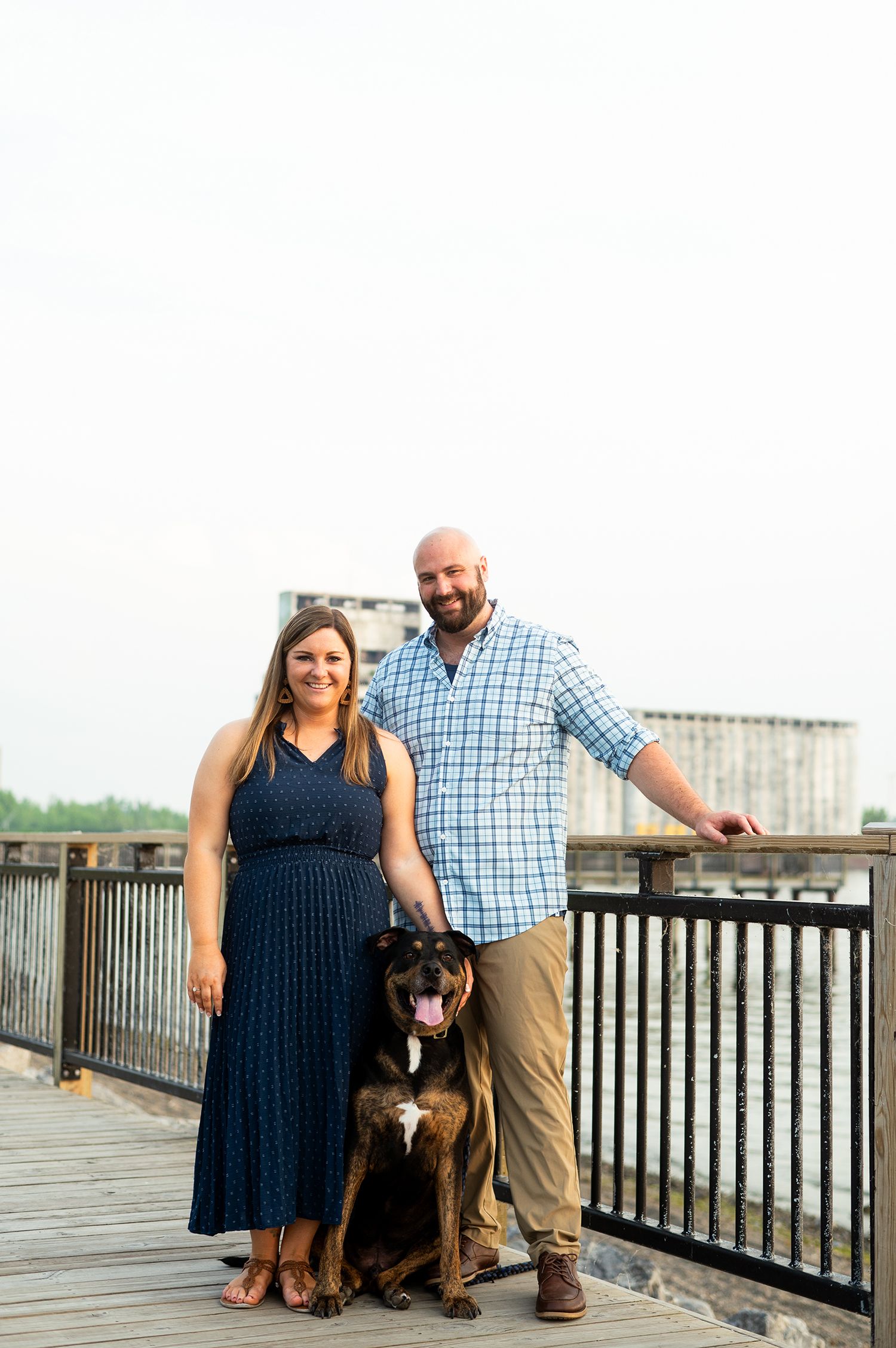 Couple in navy dress and plaid shirt posing with Rottweiler mix dog on waterfront bridge deck.